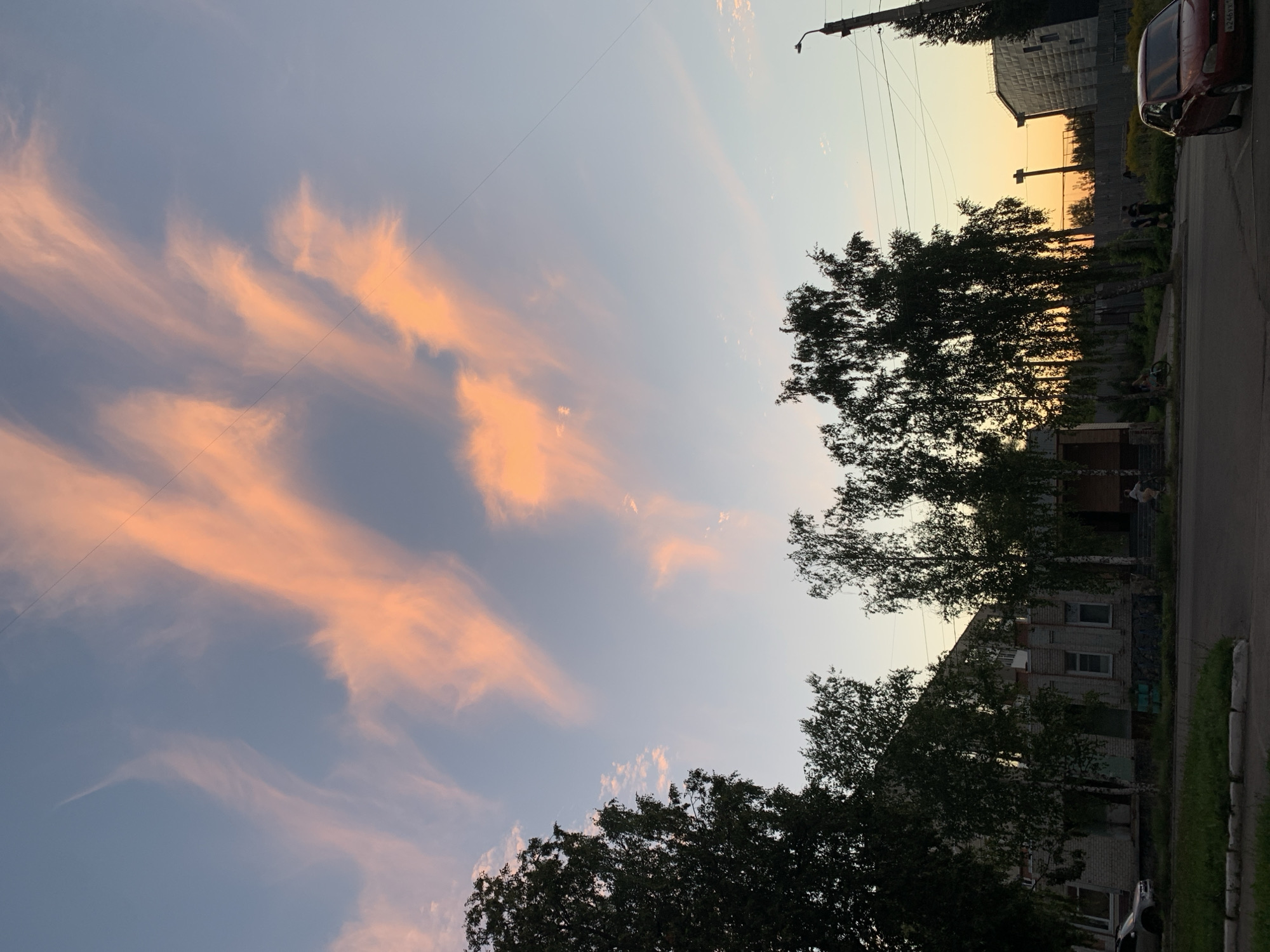 The image captures a street scene in Asha, Russia, at dusk. The sky is filled with wispy clouds tinged with orange and pink hues from the setting sun. Silhouetted against the sky are several birch trees, their foliage dark and dense. In the foreground, a street runs horizontally across the frame, bordered by a grassy area with a curb.
On the left side of the street, a two-story brick building with several windows is visible, partially obscured by trees. A silver car is parked in front of it. Further down the street, a red car is parked on the right side. Several people are present: a few individuals are walking and standing near a fence in the middle ground, and one person is visible on a bicycle in front of the brick building. A lamppost stands on the right side of the image. The overall impression is a quiet residential street as daylight fades.