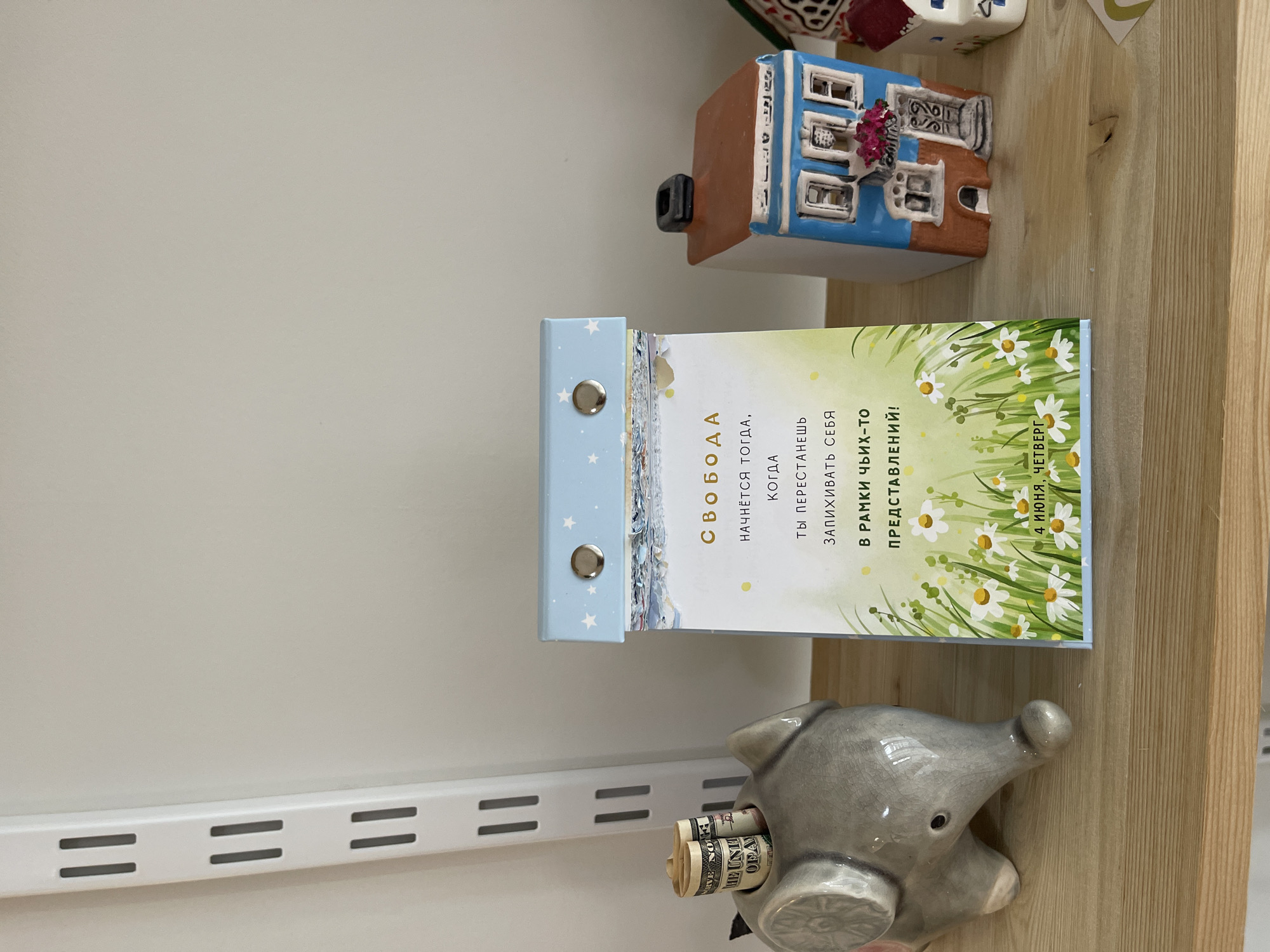 The image captures a close-up, slightly overhead view of an arrangement of objects on a light-colored wooden shelf, set against a plain white wall.
The central subject is an open daily calendar or planner, standing upright. Its visible page features a vibrant illustration of white daisies with yellow centers and green leaves against a light background, suggesting a sunny day. The top of the page has a faint illustration of what appears to be a landscape with a sky. The calendar binding is light blue with white stars, secured by two silver rings.
Prominently displayed on the calendar page is text in Russian. The large bold word at the top reads "СВОБОДА" (FREEDOM). Below it, a motivational quote states: "НАЧНЁТСЯ ТОГДА, КОГДА ТЫ ПЕРЕСТАНЕШЬ ЗАПИХИВАТЬ СЕБЯ В РАМКИ Чьих-то ПРЕДСТАВЛЕНИЙ!" (WILL BEGIN THEN, WHEN YOU STOP SQUEEZING YOURSELF INTO THE FRAMEWORK OF SOMEONE ELSE'S IDEAS!). At the bottom right, the date "4 ИЮНЯ, ЧЕТВЕРГ" (JUNE 4, THURSDAY) is visible.
To the right and slightly behind the calendar, a small, handcrafted ceramic miniature house with a brown roof and a blue facade featuring white window frames and a small red floral detail is positioned. Below the calendar, on the shelf, rests a gray ceramic elephant figure, serving as a money box. Rolled-up banknotes, appearing to be US dollars with "UNITED STATES OF AMERICA" visible on one, are tucked into the elephant's back opening. A white metal shelving track with horizontal slots is visible along the bottom left edge of the wall. The lighting is even, suggesting an indoor setting.