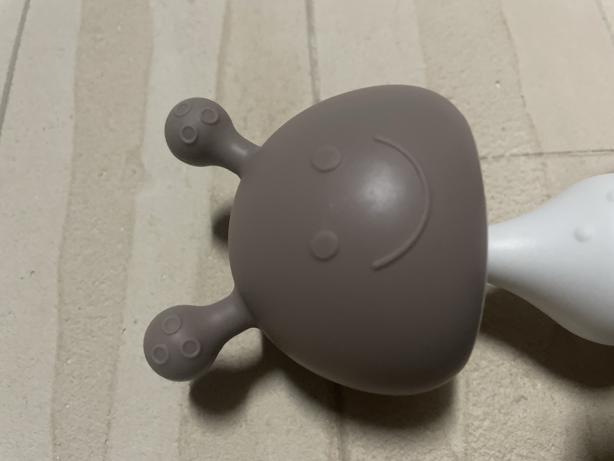 A close-up shows a gray, rubbery baby teether resembling an alien face. The "face" has two protruding arms with three circular bumps on each. A small curved line forms a smile, and two circular indentations represent eyes. The teether is connected to a white handle. The teether sits on a surface with a light beige, textured pattern resembling wood grain. There is no indication of location in Novi Sad, Serbia.