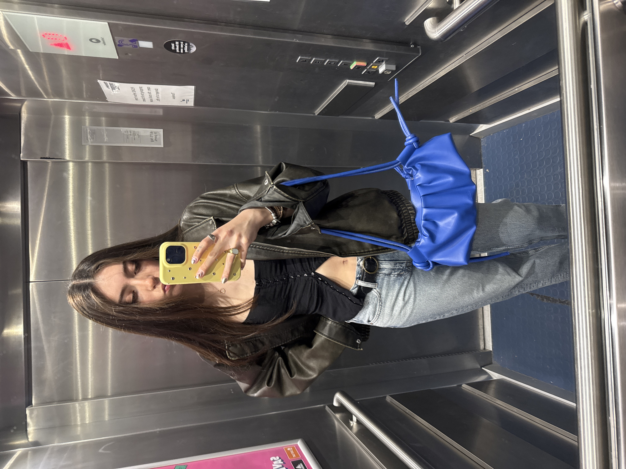 A young woman is depicted taking a mirror selfie inside an elevator. She stands in the center of the frame, holding a bright yellow smartphone with a patterned case to her face, partially obscuring her features as she looks into the phone's screen. She has long, dark hair with lighter highlights.
She is dressed in a dark brown or black leather jacket, a black ruched crop top exposing her midriff, and light blue ripped jeans with a dark belt. A vibrant royal blue, gathered-style handbag with a distinctive rope-like strap is slung over her left arm. She wears rings on her right hand, including a prominent gold-toned one, and bracelets on her left wrist. Her fingernails have white tips adorned with small black dots.
The elevator interior features reflective stainless steel walls, typical of modern lifts, which reflect the subject. The floor is covered with a dark blue, textured mat featuring circular raised patterns, and chrome handrails are visible on the sides. Above her right shoulder, an elevator control panel is visible with black buttons indicating floor numbers (1 through 6, and additional function buttons like a bell and an orange-colored one). A digital display above the buttons shows a red "1", indicating the current floor. Several white rectangular stickers with mirrored, largely illegible text, and a circular black sticker are affixed to the steel wall, including one that partially shows "E1216". The setting is an indoor location, specifically an elevator in Hull, United Kingdom.