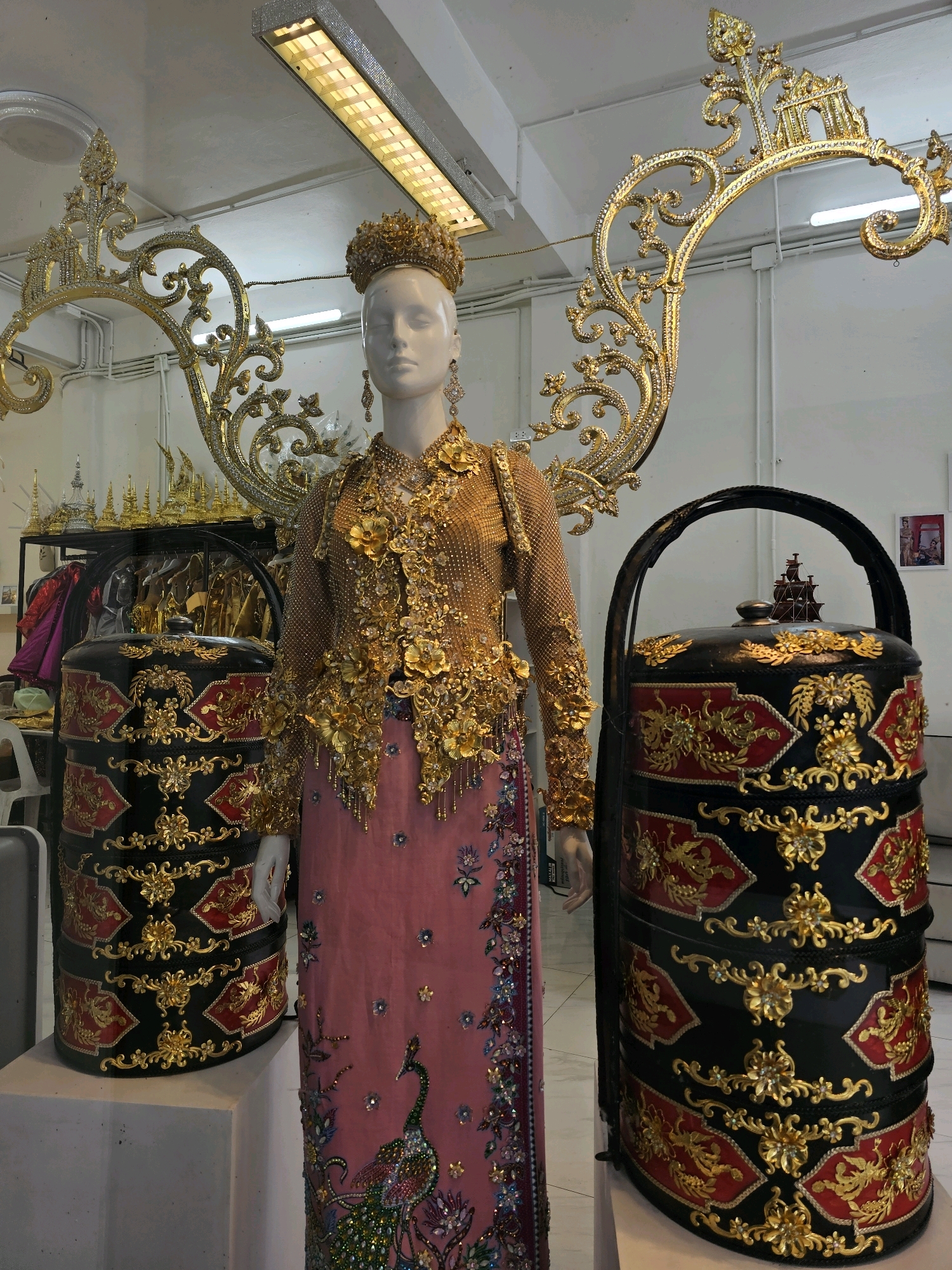 The image depicts a display of traditional Burmese attire and accessories, likely in a shop or exhibition space in Bangkok, Thailand. A mannequin, adorned in an ornate gold embroidered top and a pink skirt embellished with a peacock motif and colorful beadwork, stands at the center. The mannequin wears a golden crown and large dangling earrings. To the left and right of the mannequin are stacked, decorative containers, possibly for food or gifts, in black with intricate gold and red floral patterns. Behind the mannequin, a large, gilded, scroll-like decorative piece frames the upper body. In the background, racks of clothing and smaller decorative items are visible, suggesting a retail or cultural display setting. The overall impression is one of opulence and cultural heritage.