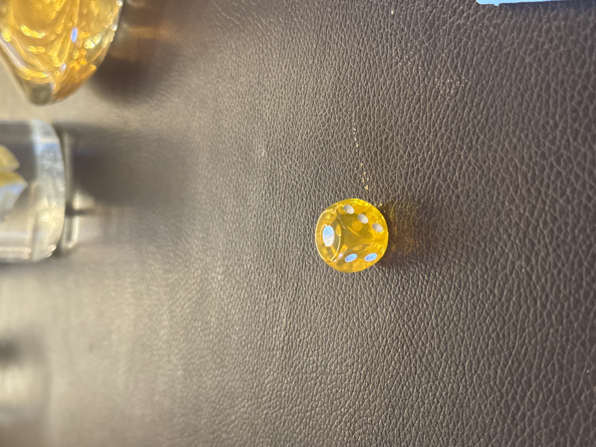 The image shows a close-up of a brown leather surface. In the upper-left corner are blurred images of glass ornaments, one appearing to be a heart shape and the other cylindrical. A small, yellow, disc-shaped object with light-colored dots is visible in the upper right. The background is plain with a dark brown textured appearance. There is no information available to link the image to North Kuta, Indonesia.
