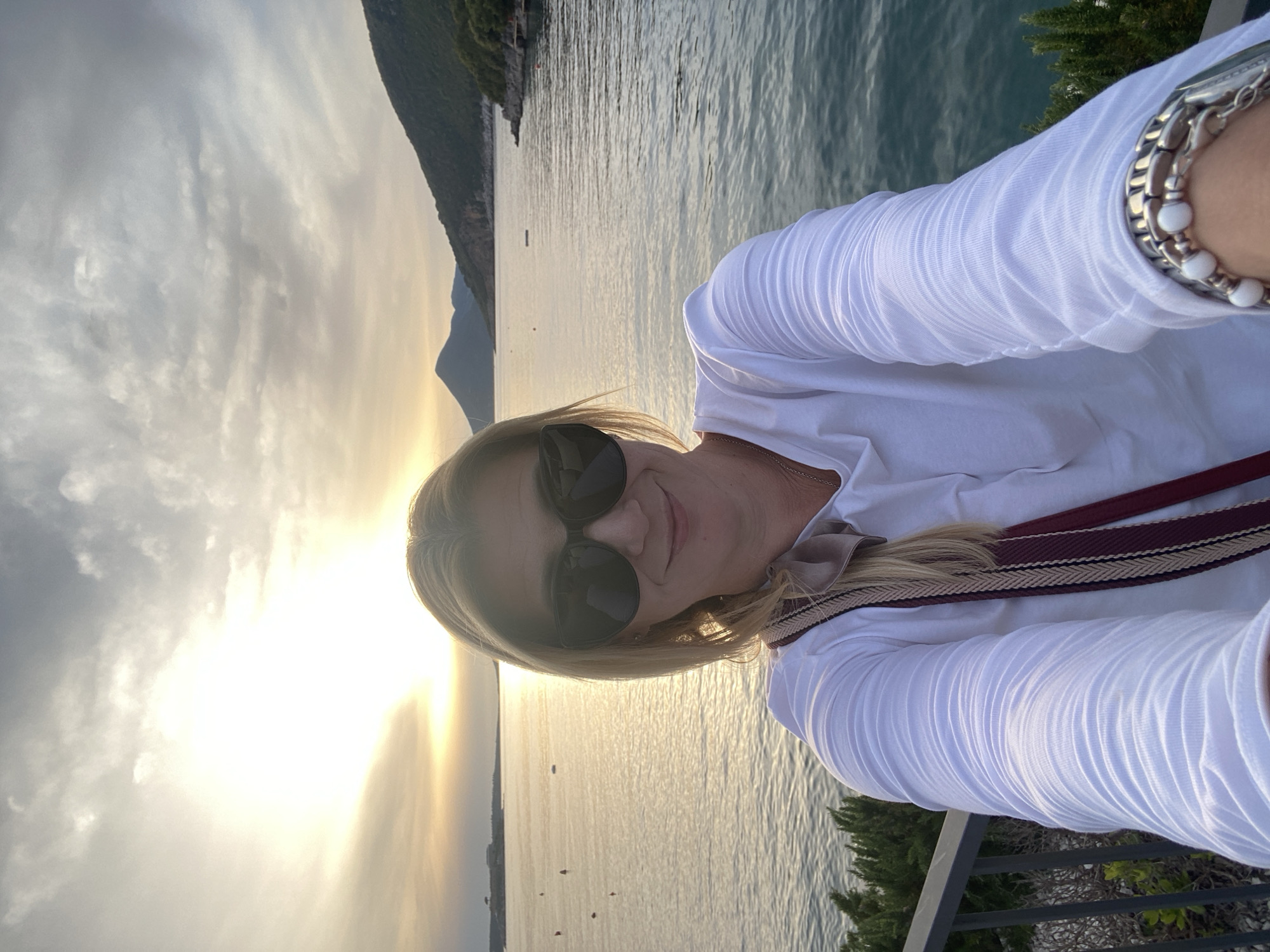 A woman with blonde hair and sunglasses is taking a selfie with a body of water in the background, likely from Luštica, Montenegro. The sun is setting, casting a golden glow on the water. There are mountains visible in the distance. The woman is wearing a white shirt and has a bracelet on her wrist. She also has a strap of some kind that is brown and white. There is a railing and some greenery visible in the foreground.