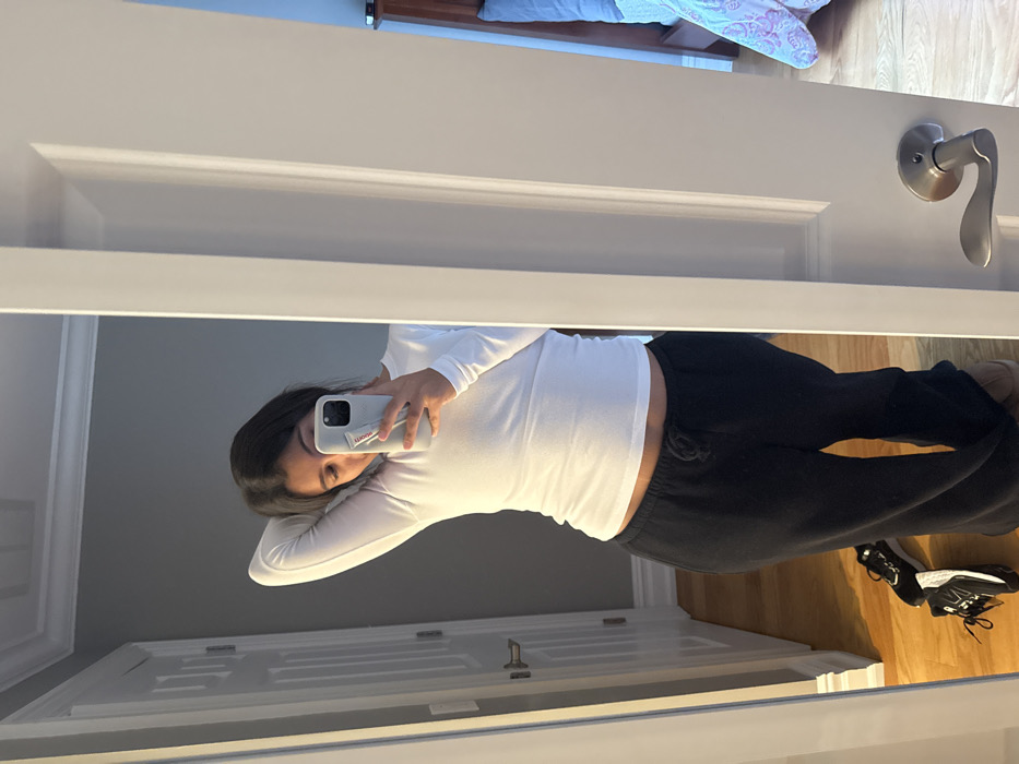 The image displays a female individual taking a mirror selfie indoors, located in a residence within Princeton, United States. The individual, possessing dark hair, is reflected from approximately the mid-thigh upward. She is wearing a white, long-sleeved shirt that exposes a portion of her abdomen, and black sweatpants with a visible drawstring. Her right hand holds a light-colored smartphone, equipped with a white grip accessory, positioned to obscure the lower part of her face. Her left arm is raised with the hand resting behind her head.
The reflection reveals a room with medium-gray walls, white crown molding, and a white paneled door featuring a silver lever handle, visible on the left side. The flooring throughout the visible areas is light-colored hardwood. Near the individual's feet, two dark-colored athletic shoes are present on the floor, one oriented on its side. To the right of the camera's perspective, through an open white door frame, another room is partially visible. This room features light blue walls, a dark brown wooden headboard, a blue pillow, and a multi-colored patterned comforter on a bed. A silver lever door handle is also visible on the white door frame in the foreground, separating the space where the photograph is being taken from the room with the bed.