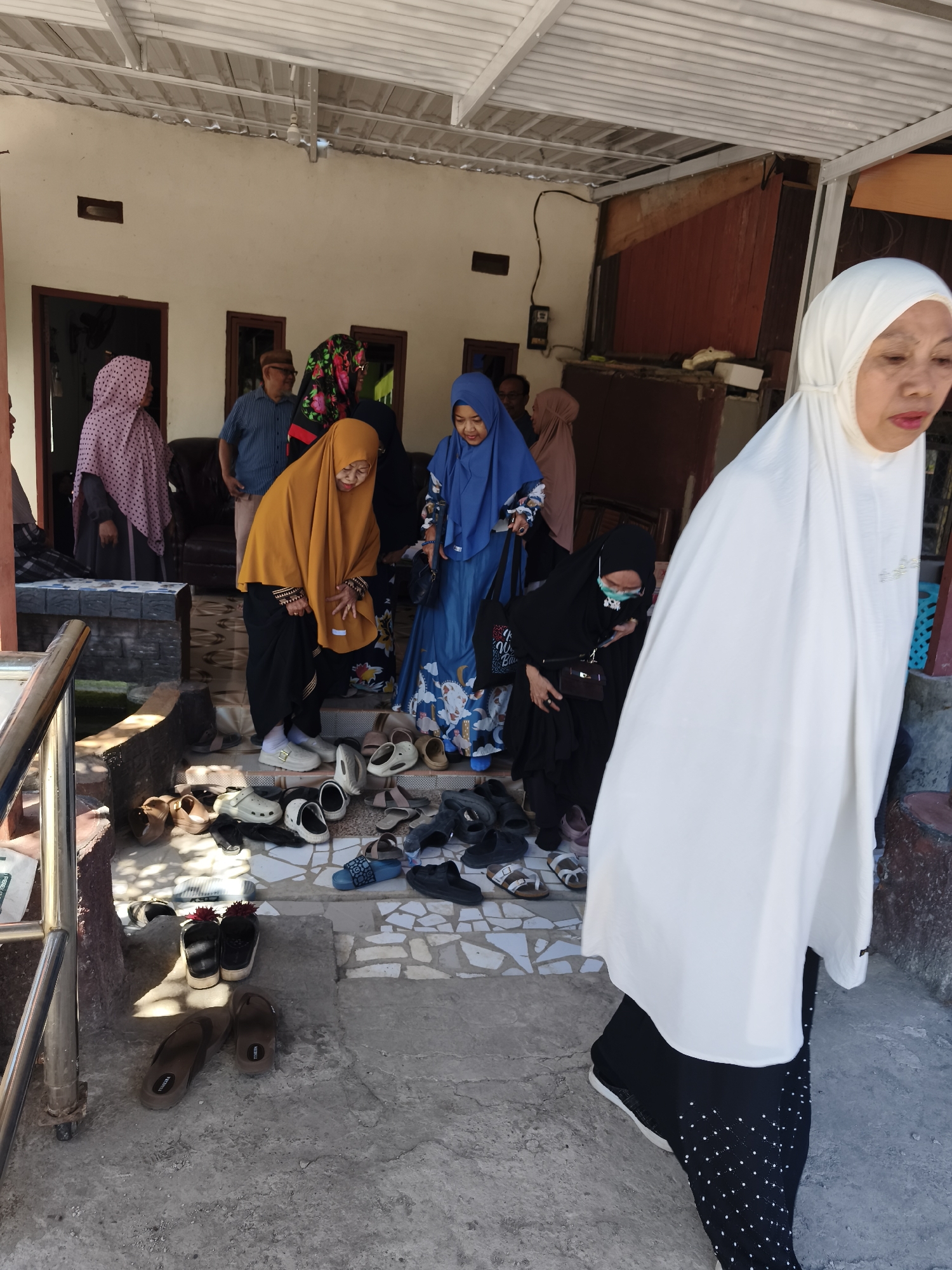 The image depicts a group of people in an outdoor setting, likely in front of a building with an open overhang. Several individuals are wearing traditional Islamic clothing, including headscarves (hijabs) and long dresses or tunics. In the foreground, a woman in a white hijab and black patterned skirt is walking towards the right of the frame. Behind her, a woman in a black hijab and glasses is looking down. Several other women are visible in the background, some wearing colorful hijabs and dresses. A man in a blue striped shirt and a dark cap is also present. The ground is paved with concrete and tile, and a collection of various shoes and sandals are arranged on steps and the ground. The architectural style suggests a residential or community building in a tropical or subtropical climate, consistent with locations in Tonasa, Indonesia. The overall scene appears to be a casual gathering or a moment of transition, possibly before or after entering a place of worship or a private residence.