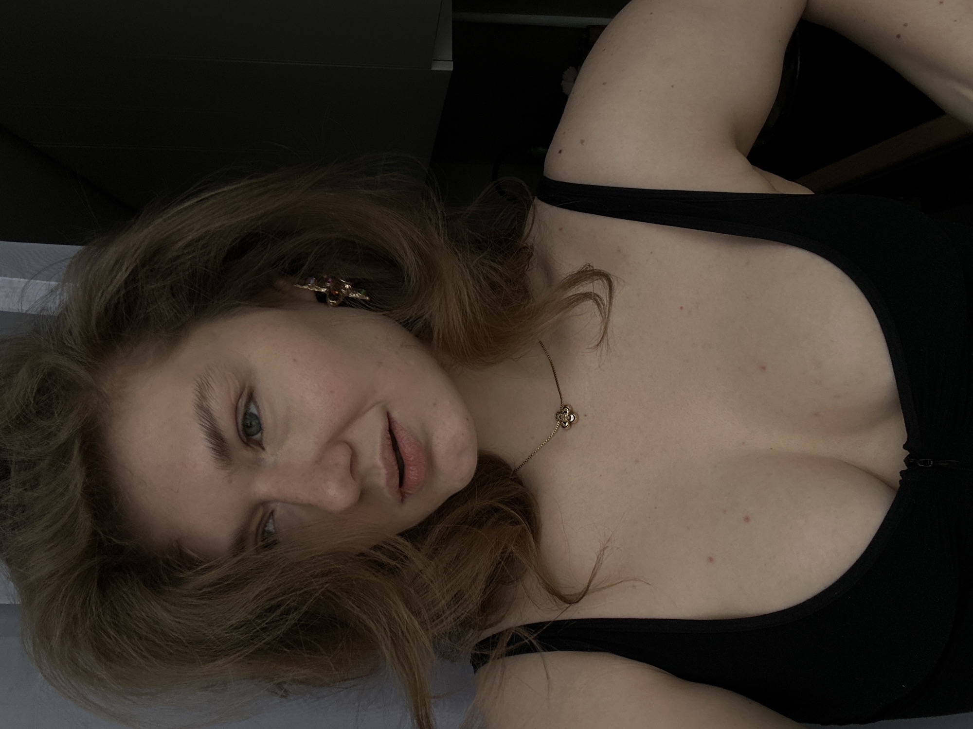 A woman with light brown hair, wearing a black tank top and a gold necklace with a four-leaf clover pendant, lies on a surface with a light background. Her head is tilted to the left, and she is looking directly at the camera with a slight smile. She is wearing earrings with a floral design. The background includes dark areas that suggest an interior space. There is no verifiable evidence of the location being Kazan, Russia in the image.