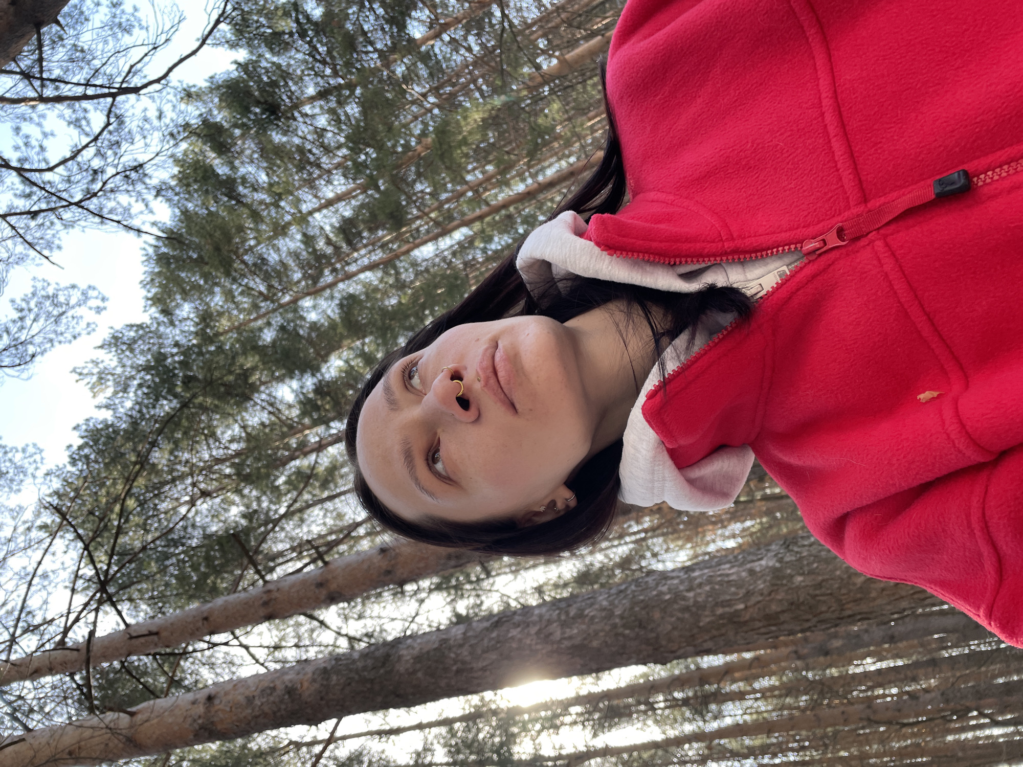 A woman with dark hair is outdoors in a wooded area. She is wearing a red fleece jacket with a white hood. The camera angle is looking up towards her face, with trees and sky in the background. The location cannot be determined with the available information.