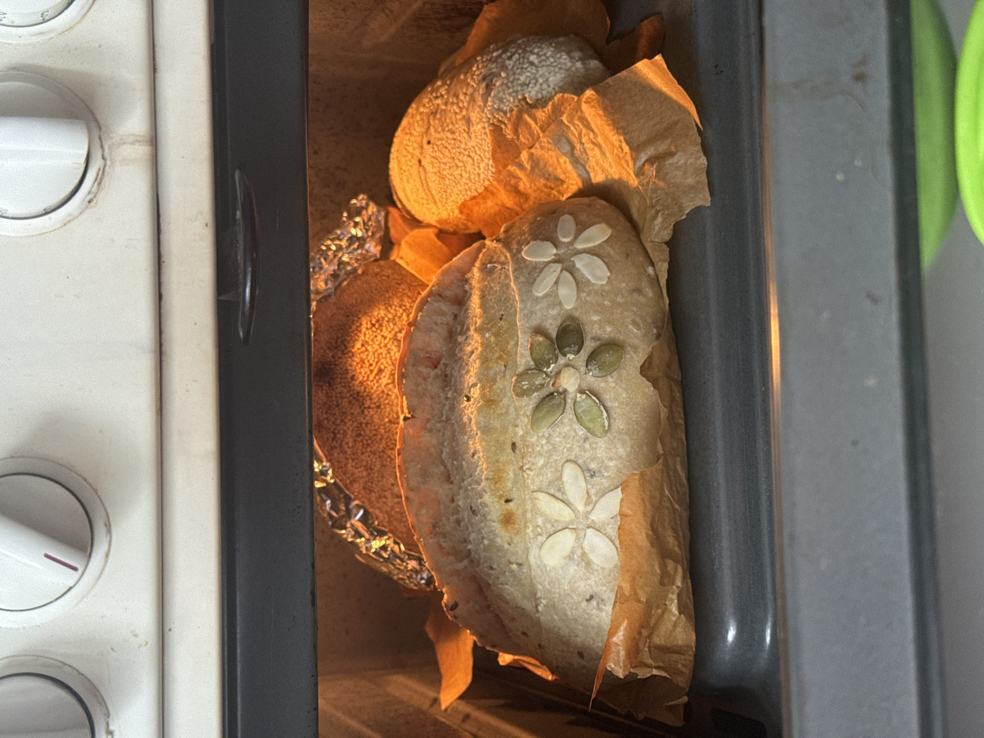 The image shows three loaves of bread baking inside an oven. The bread is resting on baking paper inside a metal tray. One loaf is decorated with seeds arranged in a flower pattern, another appears to be covered in sesame seeds, and the third is covered with other seeds. Aluminum foil is placed to protect the baking paper. The image also shows the control panel of the oven with dials and a portion of the oven door and its handle. There is nothing to indicate that this scene is taking place in the city of Высокий, Russia.