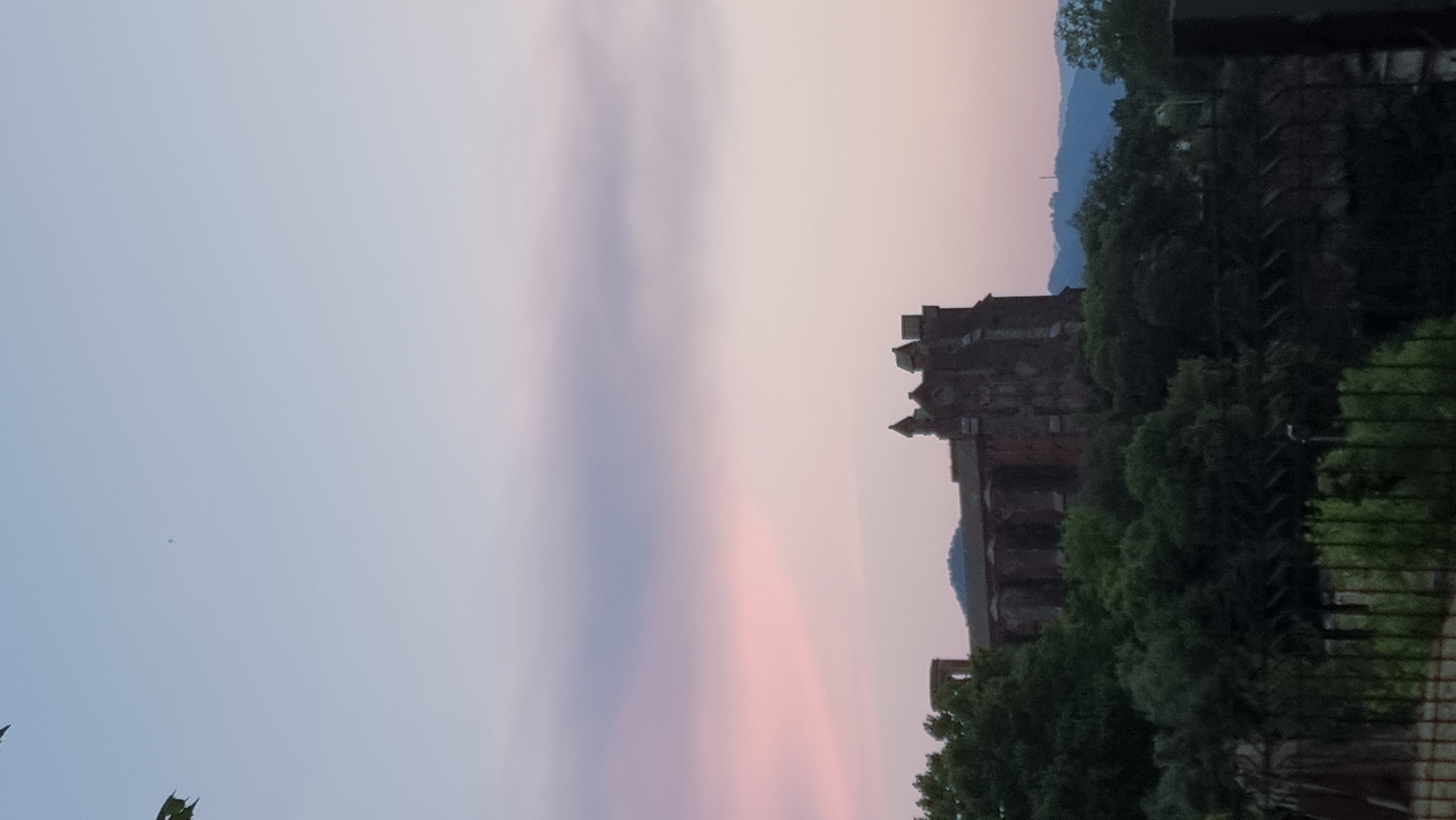 The image depicts a large, old stone building with multiple towers, possibly a church or a historic structure, nestled among lush green trees. The building appears to be made of dark stone, with intricate architectural details visible on its facade. The sky above is a soft gradient of light blue and pink, suggesting it might be either sunrise or sunset. In the distance, rolling hills or mountains can be seen, their forms softened by the atmospheric conditions. In the foreground, there is a dark, blurred area that could be a fence or railing, with some vegetation beneath it. The scene conveys a sense of tranquility and historical presence. The building is identified as part of the municipality of Sant Joan Les Fonts, Spain.
