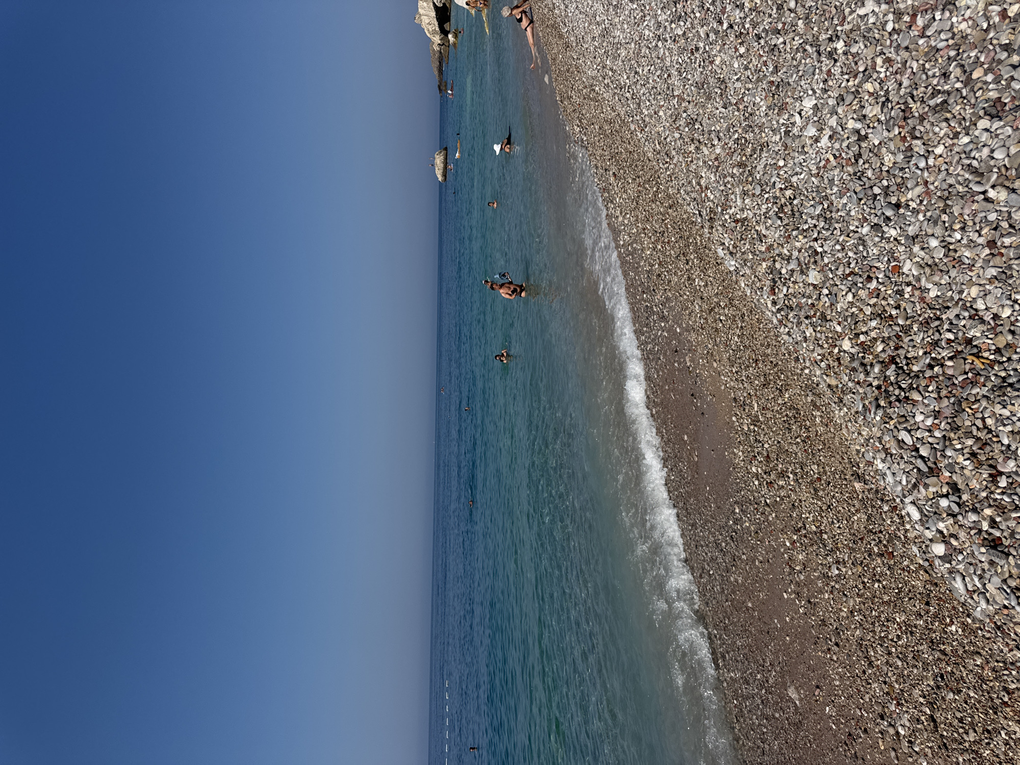 The image shows a pebble beach with clear turquoise water at the city of Mirište, Montenegro. The sky is a bright, clear blue, suggesting a sunny day. The water is calm with gentle waves lapping at the shore. Several people are visible in the water, swimming or wading, and a few are sunbathing on the pebble beach. A rocky outcrop with some vegetation is visible in the distance on the right side of the image, and a similar, smaller rock formation is in the middle distance of the sea. The beach itself is covered in smooth, multicolored pebbles of varying sizes.