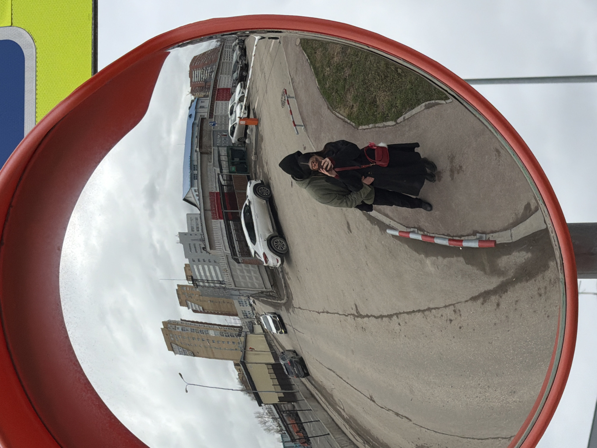 Reflection in a convex safety mirror shows two people standing close together on a sidewalk, the man in a green jacket with his arm around a woman in a black coat, a red purse, and a black hood, with the woman holding a phone. The sidewalk borders a road with parked cars and apartment buildings. The city is Perm, Russia. The weather appears overcast.