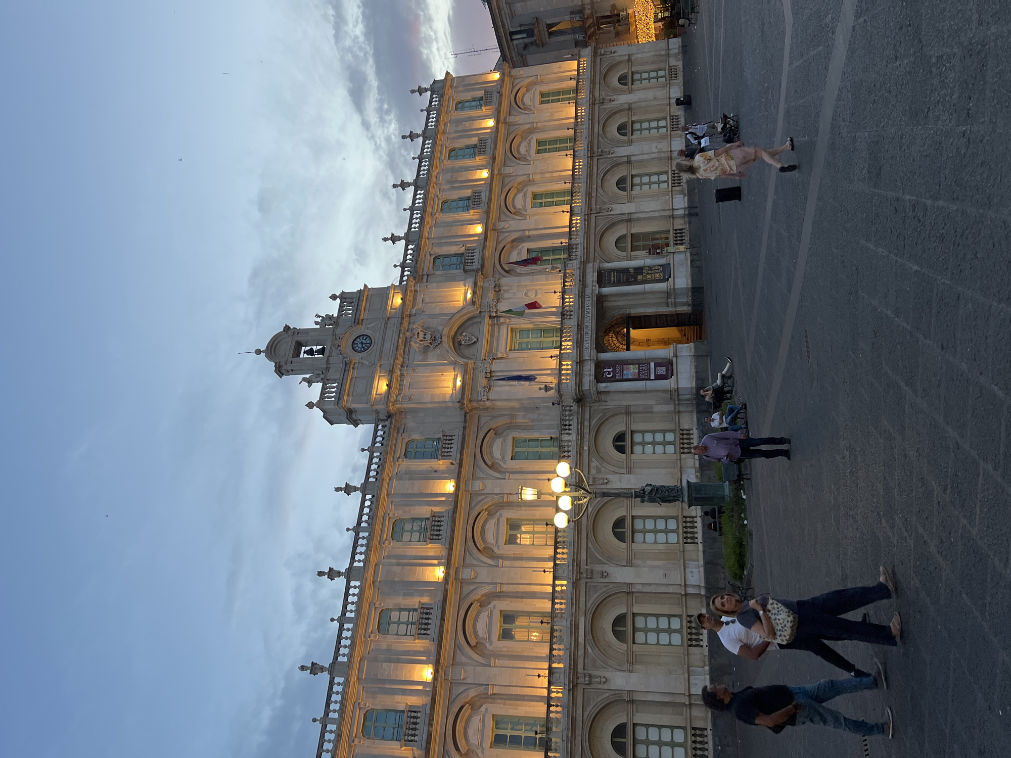 The image depicts Piazza Università in Catania, Italy, at dusk. The main subject is the historic Palazzo dell'Università, a grand Baroque building illuminated by warm yellow lights. Its facade features multiple stories with arched windows and decorative elements. A bell tower with a clock is visible at the top center of the building.
In the foreground, people are seen walking across the stone-paved piazza. To the left, three individuals are walking together: a man in a black t-shirt and jeans, a man in a white t-shirt and dark pants, and a woman in dark pants and a printed bag. Further to the right, a man in a purple shirt stands looking towards the right. Several other people are visible in the middle ground, some sitting on benches.
Streetlights and decorative lights, possibly from a nearby building or event, add to the ambient illumination of the scene. The sky is a soft blue with scattered clouds, indicating the transition from day to night. The overall atmosphere is one of a public square bustling with evening activity.