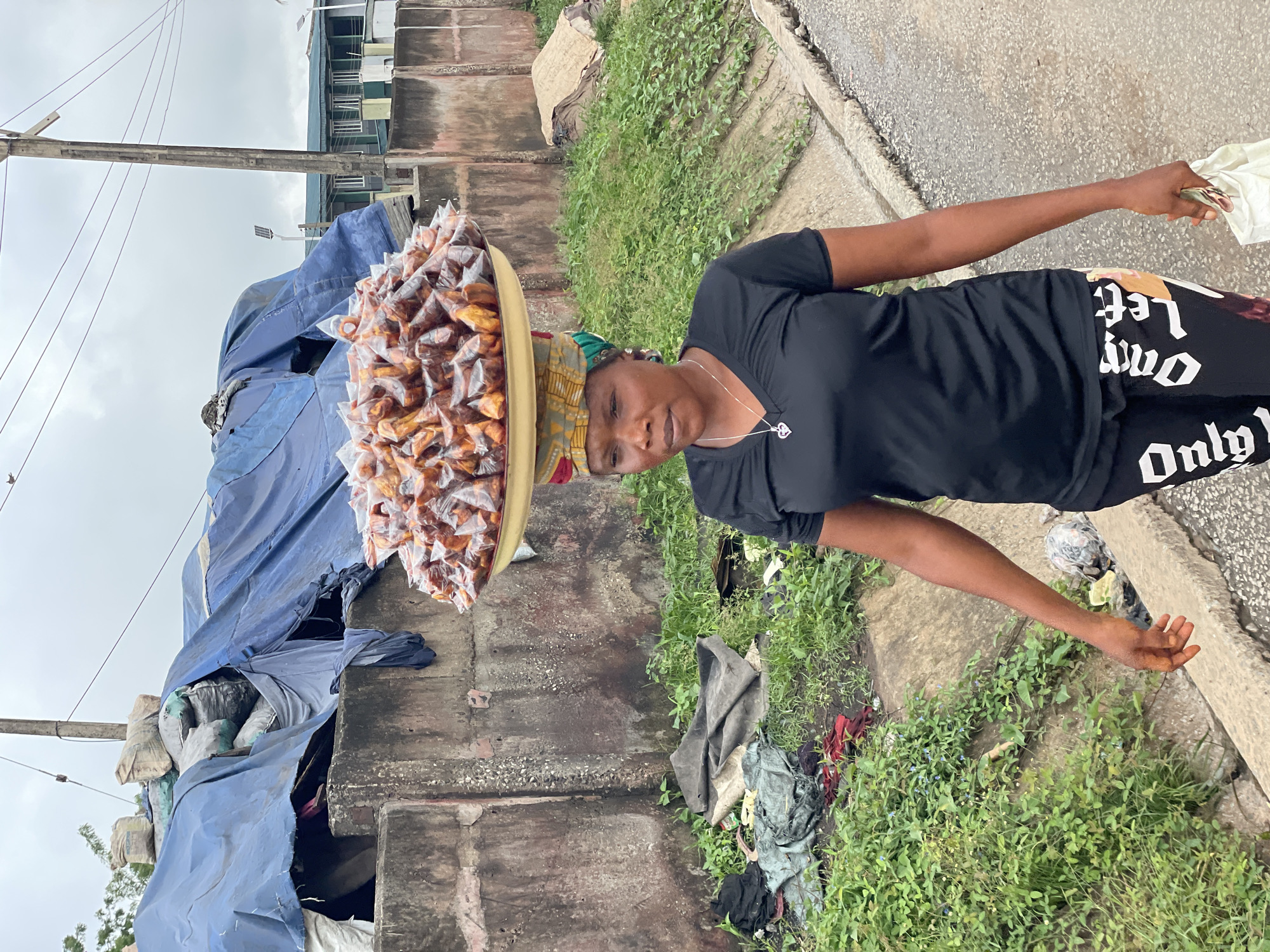 A woman walks down a street in Fegge, Nigeria, carrying a large tray of food items on her head. The food items are individually wrapped in clear plastic bags and appear to be fried plantains. The woman is wearing a black t-shirt, black leggings with white lettering, and a colorful headscarf. She is walking on a paved road next to a drainage ditch filled with green vegetation. In the background, there is a concrete wall, some tarpaulin-covered materials, and buildings. The sky is overcast.