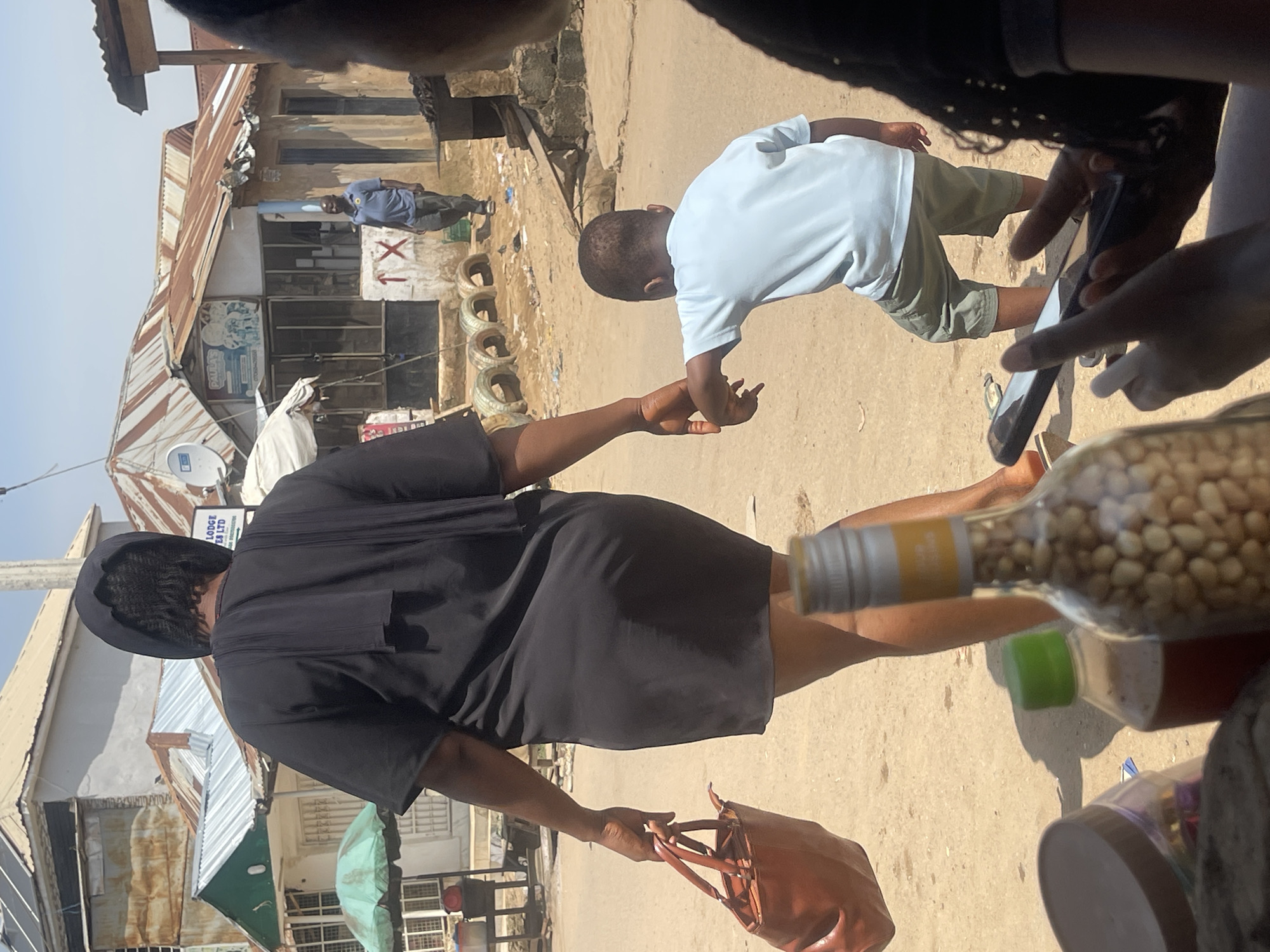 The image depicts a street scene in Abuja, Nigeria, likely a market or a residential area with shops. In the foreground, from left to right, are parts of bottles containing food items, a brown leather handbag held by a woman, and a young child. The woman, wearing a black dress and headscarf, holds the child's hand. The child, wearing a light blue t-shirt and green shorts, is facing away from the camera. A partially visible person, likely also female, is in the extreme right foreground, holding a smartphone. The background shows a row of shops with corrugated iron roofs, some appearing aged. A man in a blue shirt and grey trousers stands near one of the shops. The ground is dusty and appears to be a dirt road. Several discarded tires are visible in front of one of the shops, and some trash is scattered around. The overall environment suggests a typical urban street in Nigeria, possibly on a sunny day given the strong shadows.