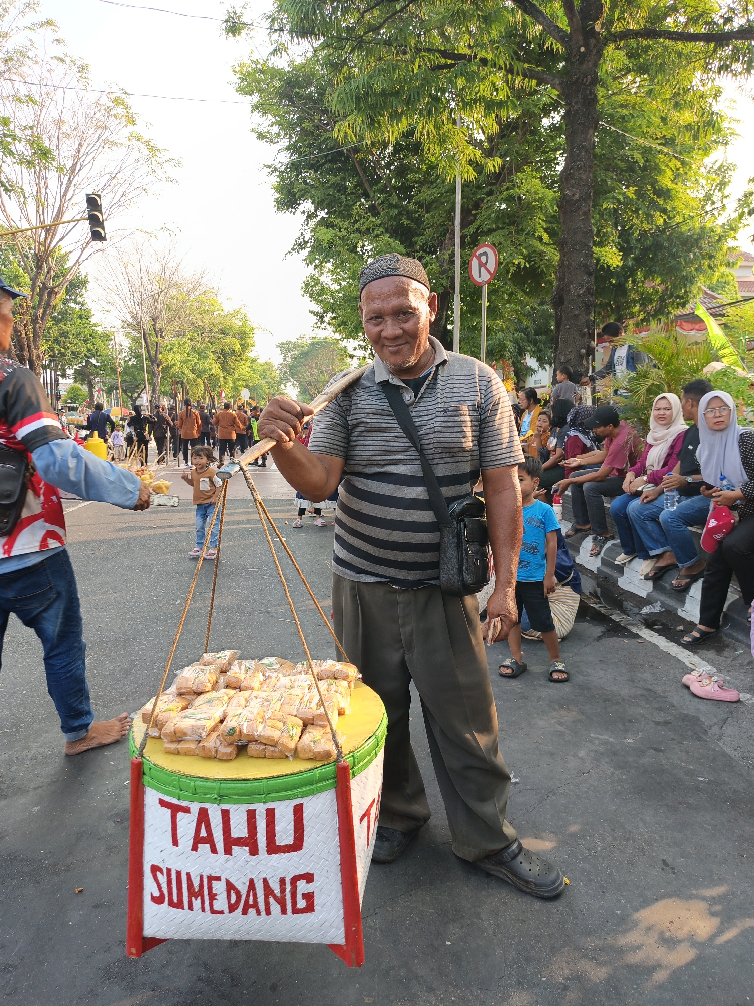An outdoor medium shot captures a middle-aged Indonesian man in a traditional cap and striped t-shirt, carrying a basket of "Tahu Sumedang" (fried tofu) on his shoulder in Klaten, Indonesia. The basket, painted white with red trim and yellow accents, displays the product's name in red lettering. The man faces the camera, smiling, with a black sling bag over his shoulder. The background shows a street scene with trees, other pedestrians, and traffic lights. The setting appears to be a busy public area, possibly a market or a street festival, given the presence of various individuals and colorful decorations in the distance. The lighting suggests it is daytime, likely late afternoon or early evening due to the warm, soft sunlight.