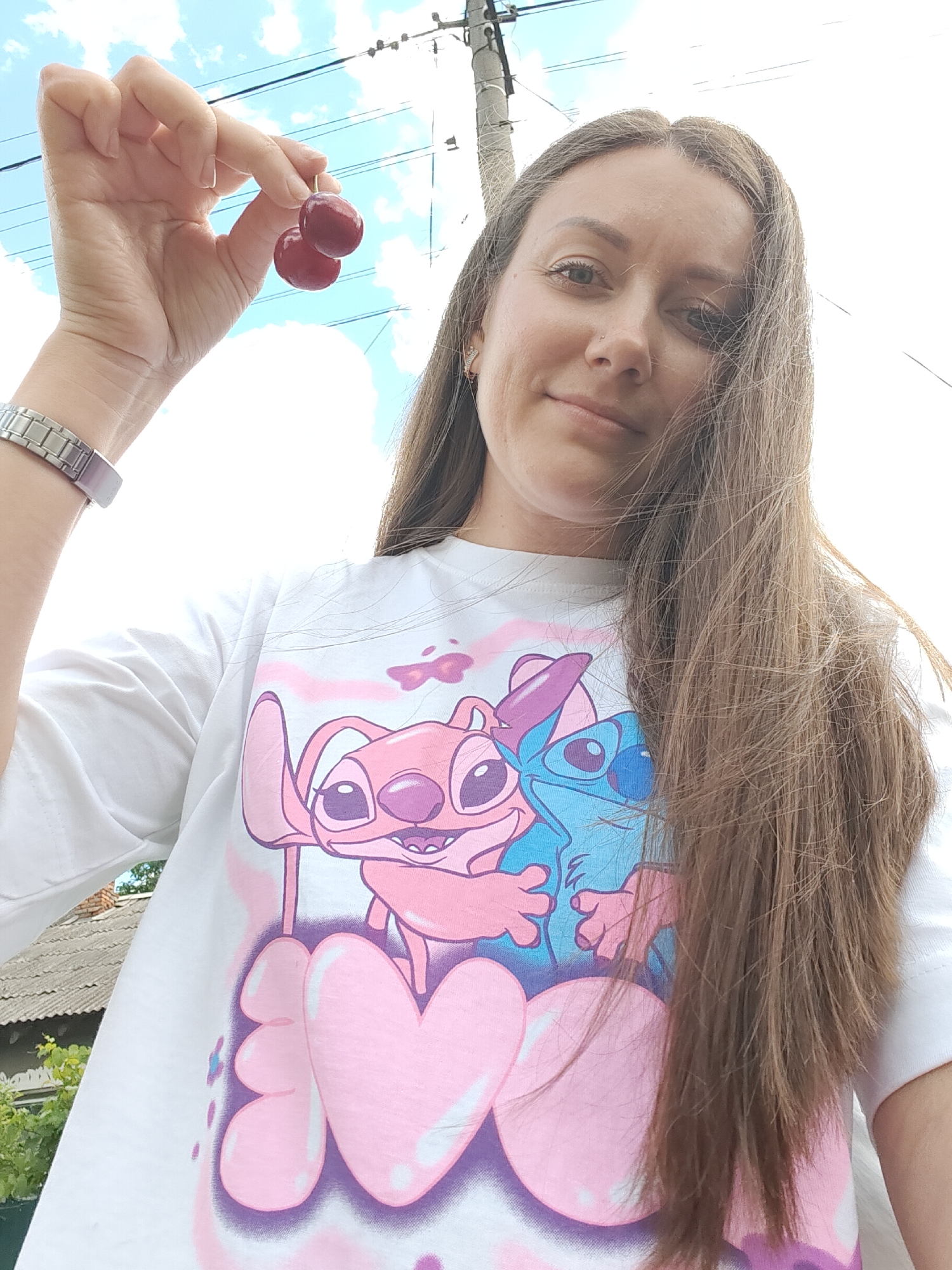 A young woman is holding two cherries between her thumb and forefinger, presented up towards the sky. She is wearing a white t-shirt with a cartoon design of two characters, one pink and one blue. The woman has long, light brown hair and is looking towards the camera with a slight smile. Her left arm and hand are visible, wearing a silver watch. The background shows a bright sky with some clouds and overhead power lines, and a glimpse of a rustic roof and greenery to the left. The scene appears to be outdoors, possibly in a residential area.