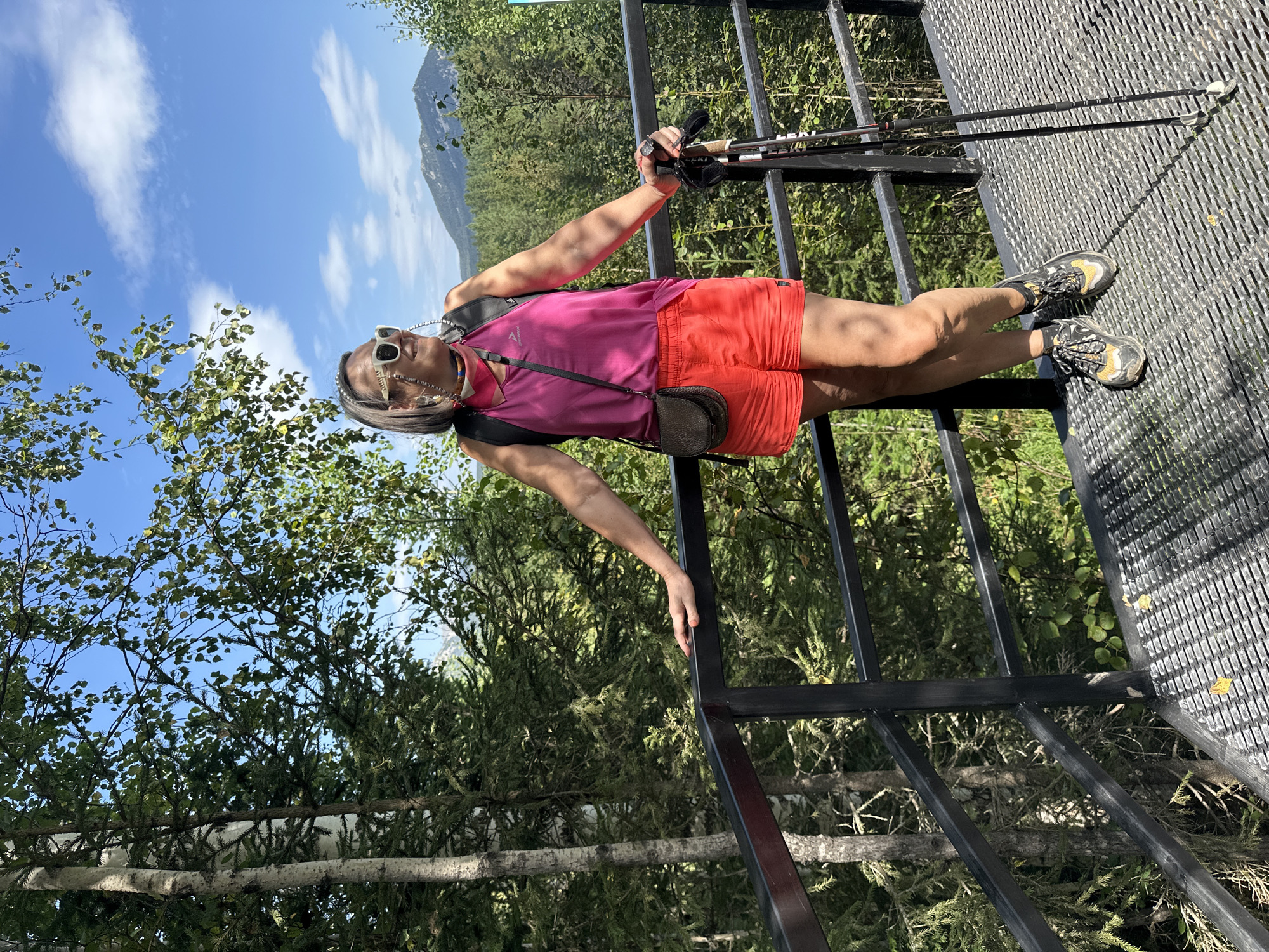 A woman stands on a metal platform outdoors, leaning on a black metal railing. She is wearing a pink tank top, red shorts, sunglasses, and hiking boots, and is holding two hiking poles. The background shows a forest with tall trees and a blue sky with clouds. In the distance, a mountain is visible. The scene appears to be in a natural, possibly mountainous, setting.