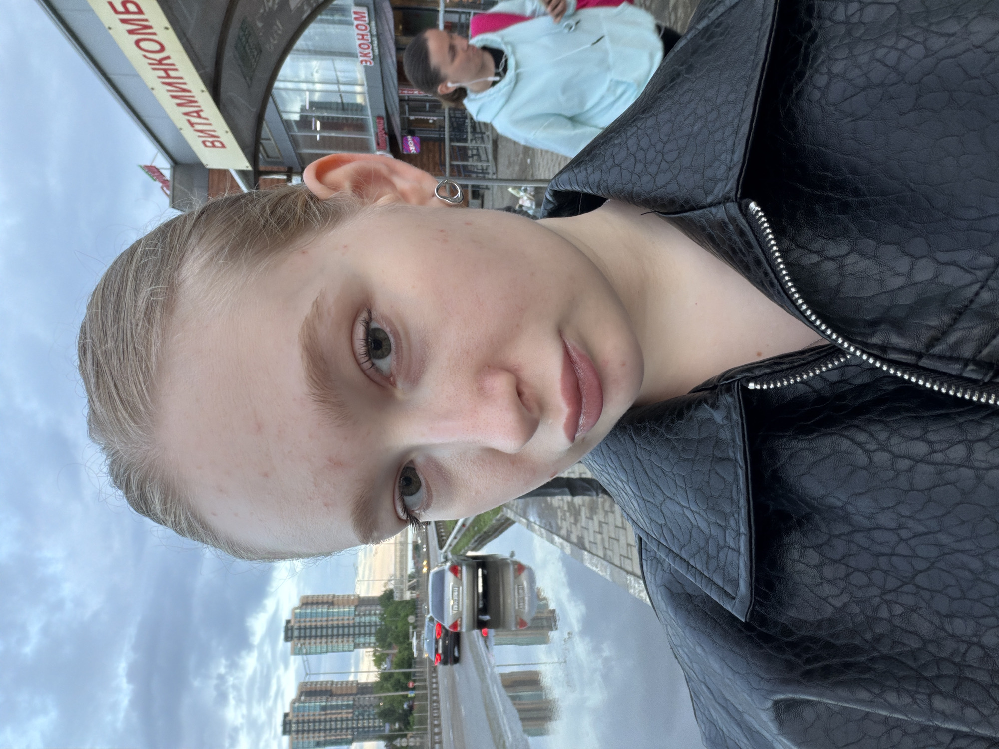 A selfie of a young woman with fair skin and light-colored hair, wearing a black leather jacket. She is near a street with visible traffic. Reflected in a wet surface, likely the pavement, are modern residential buildings. A sign reading "ВИТАМИНКОМБ" is visible in the background. Another person wearing a light blue hoodie is also in the shot. Given the Russian signage, this image appears to be taken in the city of Beryozovsky, Russia.
