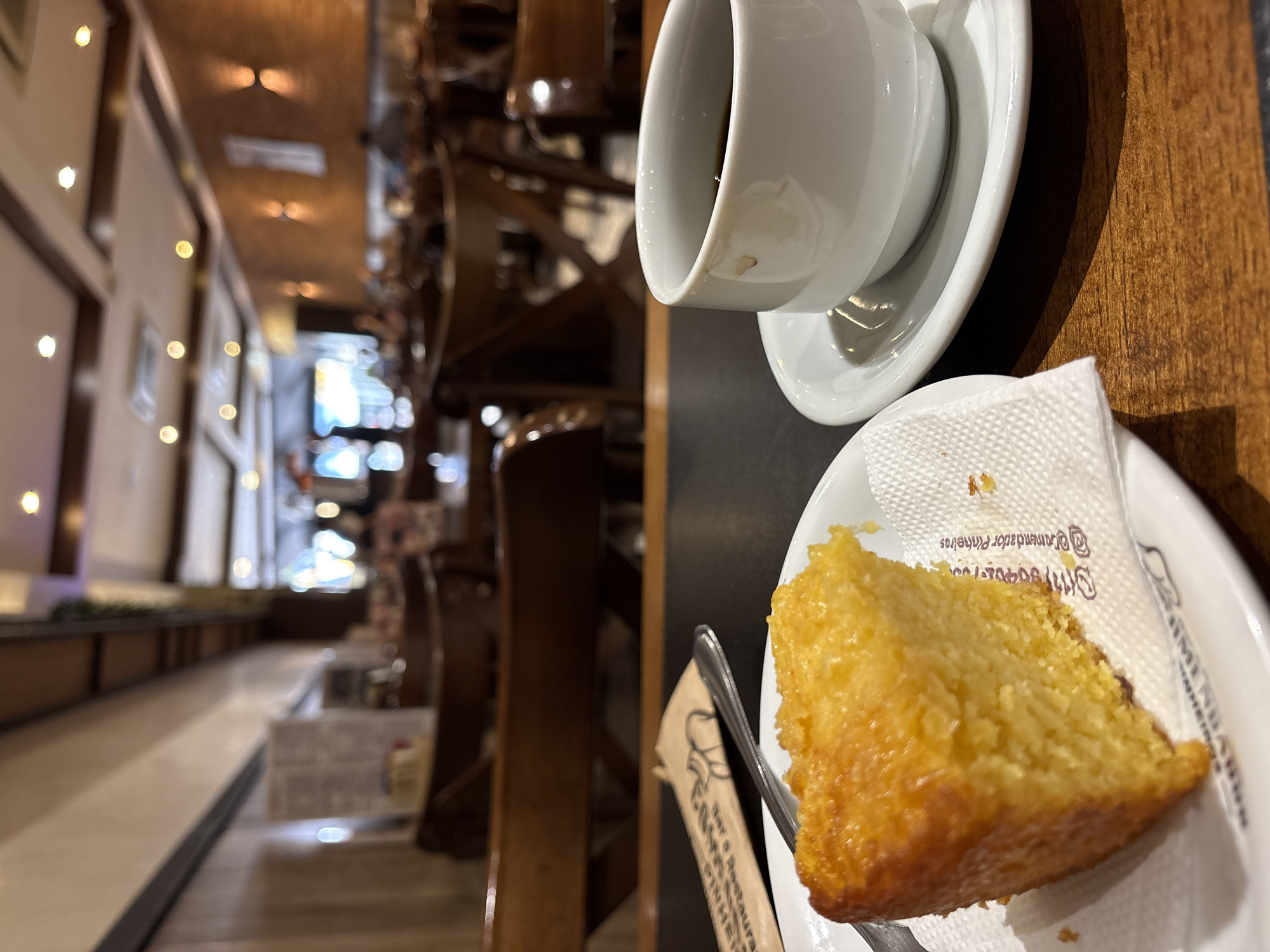 A close-up shot shows a slice of yellow cake on a white plate next to a white cup of coffee on a saucer. The cake is moist and has a slightly golden-brown top. The coffee is dark and appears to be black. A white paper napkin with some text and an illustration is placed under the cake. The setting appears to be a cafe with wooden tables and chairs visible in the background. The cafe has warm lighting and brick walls, suggesting a cozy atmosphere. The blurry background indicates that the focus is on the food and drink in the foreground. The overall scene captures a moment of enjoying a simple yet satisfying treat.
