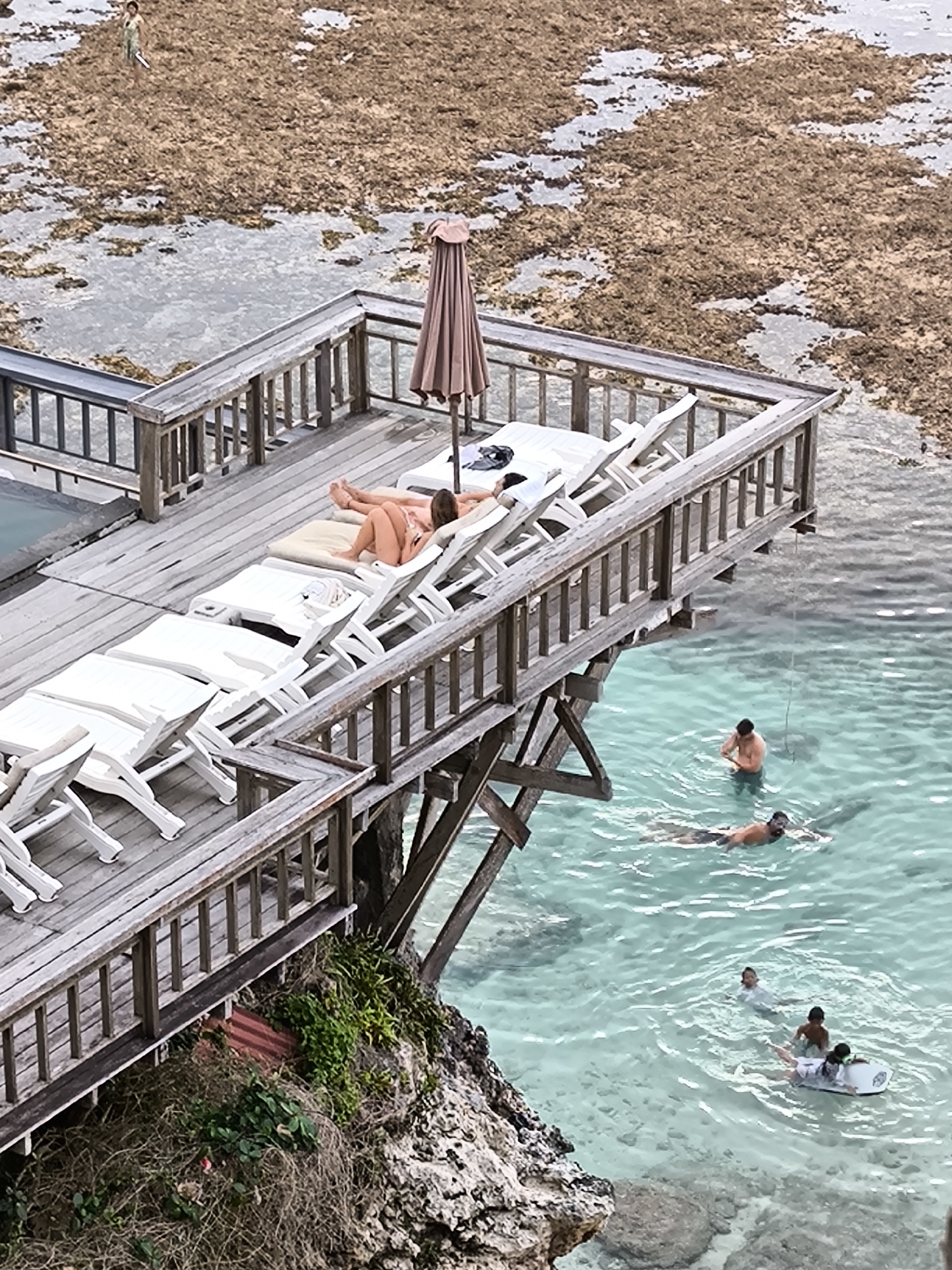 The image shows a wooden deck perched on a rocky cliff overlooking turquoise water, likely in Uluwatu, Indonesia. On the deck, there are several white lounge chairs, some empty and some stacked. A woman is reclining on one of the chairs. An open umbrella stands nearby. In the water below, several people are swimming and snorkeling. One individual is on a surfboard. The rocky area near the deck has some vegetation growing on it. In the background, the visible terrain has a mix of water and land covered with what appears to be vegetation or algae. A person is seen walking in this background area.