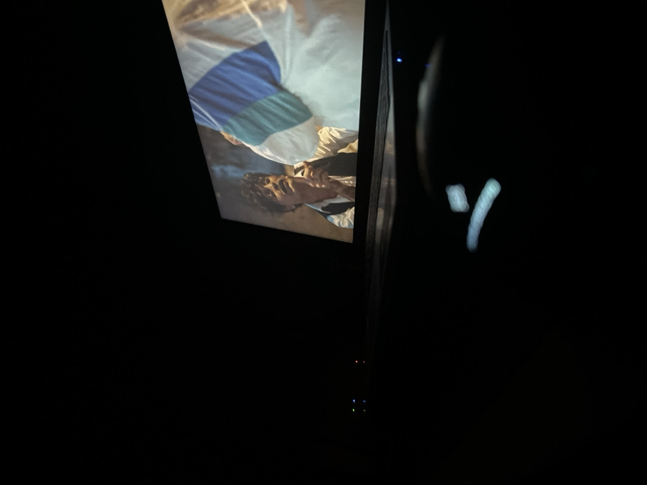 In a dimly lit room, a vertical rectangular screen displays a scene with a person lying down, wearing a white shirt with blue and green stripes. The person's face is visible. To the right of the screen, there is a blurred, pale, "Y" shaped light. Further down, near the bottom of the frame, several small LED lights are visible, indicating an electronic device. The rest of the image is dark.