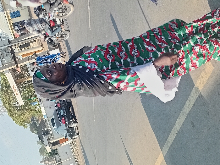 An older woman wearing a green, red, and white patterned dress and black headscarf stands on a sunlit street. A white shirt is visible under the dress's sleeves. Cars and motorcycles populate the background, along with buildings and trees. A sign for "MUHARN" is also visible. The woman appears to be looking upwards. The scene is captured from a slightly elevated angle.