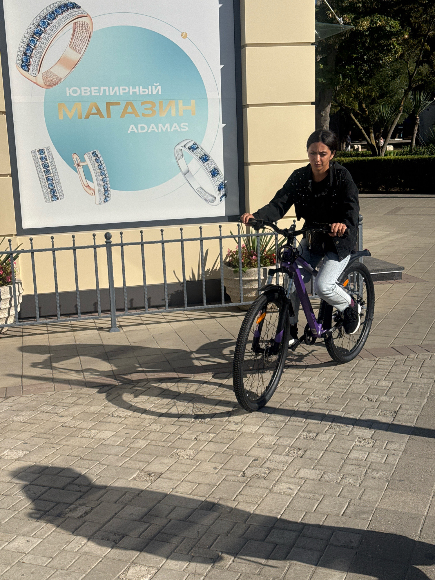 A woman rides a purple bicycle on a paved walkway in front of a building with a large advertisement featuring jewelry. The woman, wearing a black jacket and light-colored pants, is positioned in the center of the frame. The advertisement, displayed on a light blue circular background, is for a jewelry store named "ADAMAS" and shows images of rings and earrings. Behind the woman, a metal fence separates the walkway from the building's facade. The ground is paved with rectangular bricks, and the sunlight casts long shadows of the woman and her bicycle onto the walkway. The environment appears to be an outdoor urban setting.