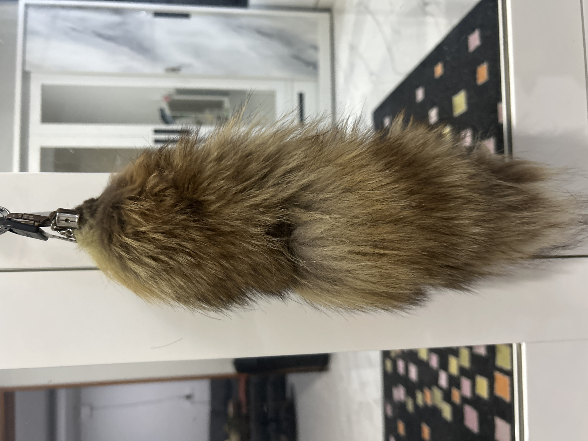 A fluffy, brown, and tan fur keychain is hanging from a metal clasp. The keychain is suspended in front of a white, reflective surface, possibly a closet door or a mirror. To the left of the keychain, a reflection shows a dimly lit room with furniture. To the right, the reflection reveals a modern interior with white closet doors and patterned floor mats. The overall scene appears to be indoors, likely within a residential or commercial space. The location in Sukawati, Indonesia is not visually discernible from the provided image.
