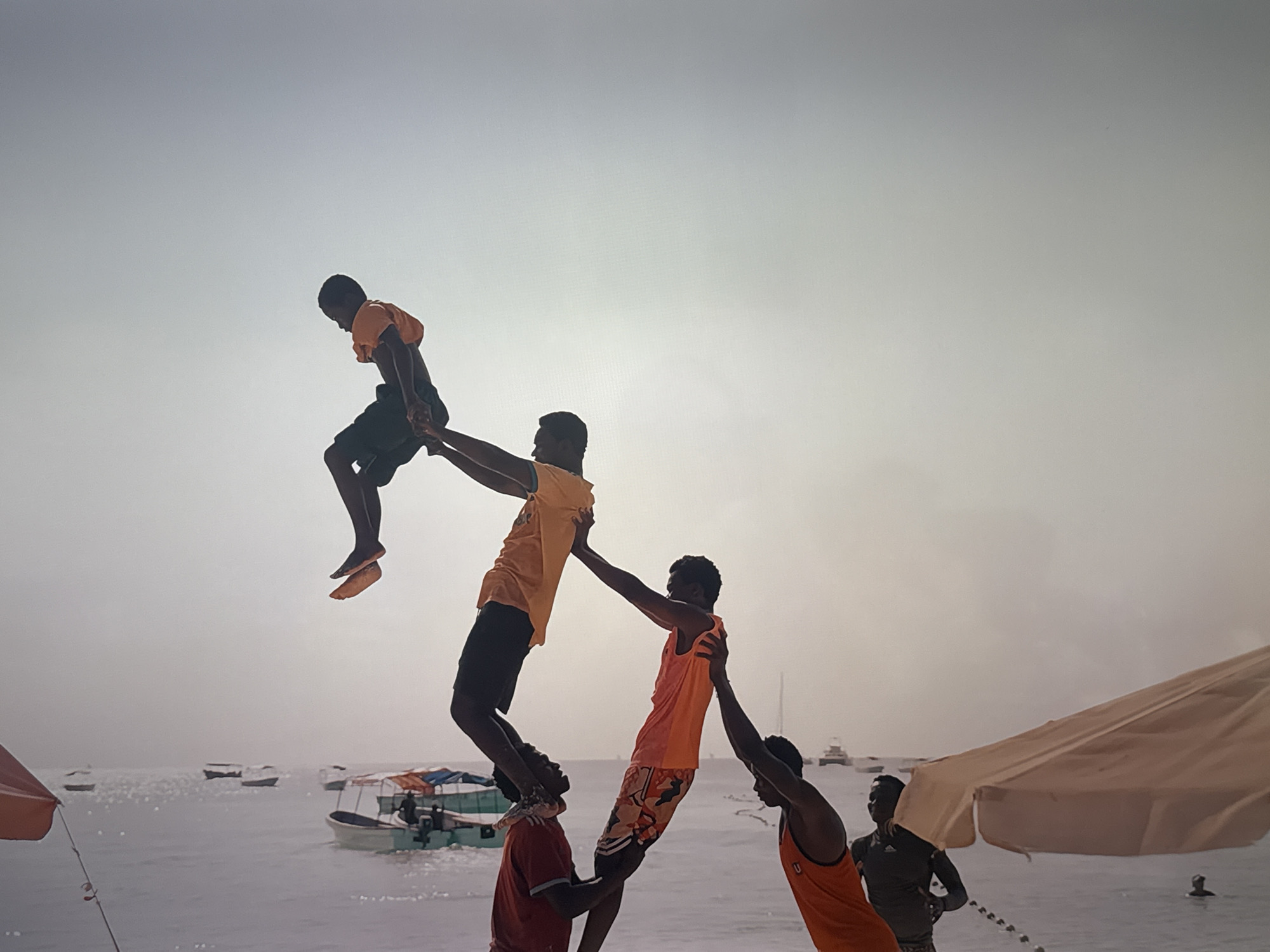 In an outdoor scene with a hazy sky, a group of five men are performing an acrobatic human pyramid on what appears to be a beach or pier. The man at the top, wearing an orange t-shirt and dark shorts, is suspended mid-air, being held by two other men. The man below him, in an orange tank top and patterned shorts, is supported by another man in a red t-shirt. The lowest man in the formation is holding the man above him. In the background, several boats are visible on the water, and a large beach umbrella is on the right side of the frame. The overall environment suggests a coastal location.