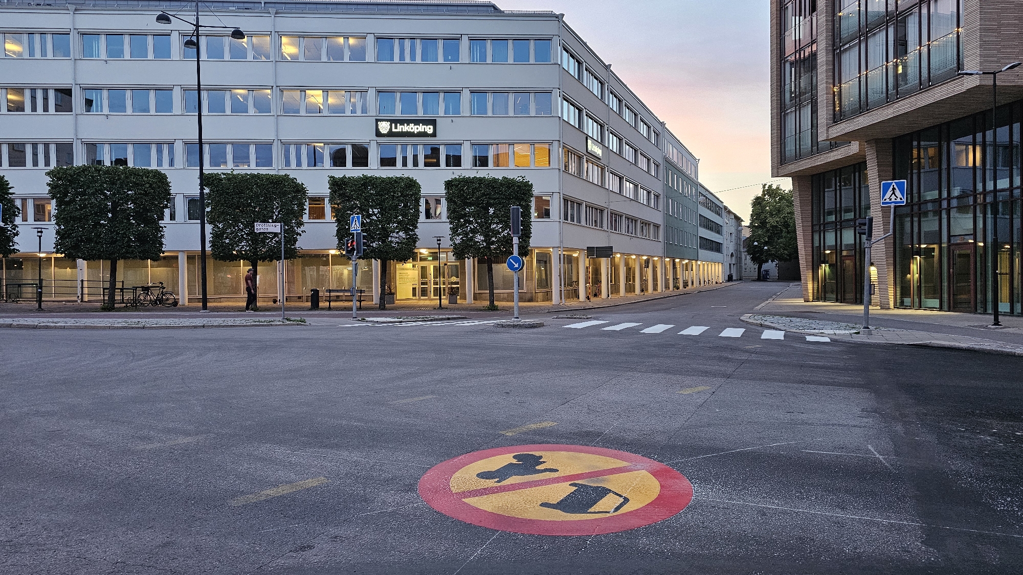 The image captures an intersection in Linköping, Sweden, at dusk. In the foreground, a red and yellow circular road sign depicts a silhouette of a person with a dog on a leash, with a red diagonal line through it, indicating no dogs allowed. White crosswalk lines are visible on the asphalt. To the left, a modern, light-colored building with numerous windows and a "Linköping" logo is prominent. Several evenly-spaced, square-shaped trees with dark foliage line the sidewalk in front of this building. A single person is walking on the sidewalk, and a bicycle is parked. To the right, a contemporary building with a glass facade and a wooden exterior wraps around the corner, its windows illuminated. A blue pedestrian crossing sign is mounted on a pole. The sky shows a gradient of soft pink and blue hues, typical of twilight.