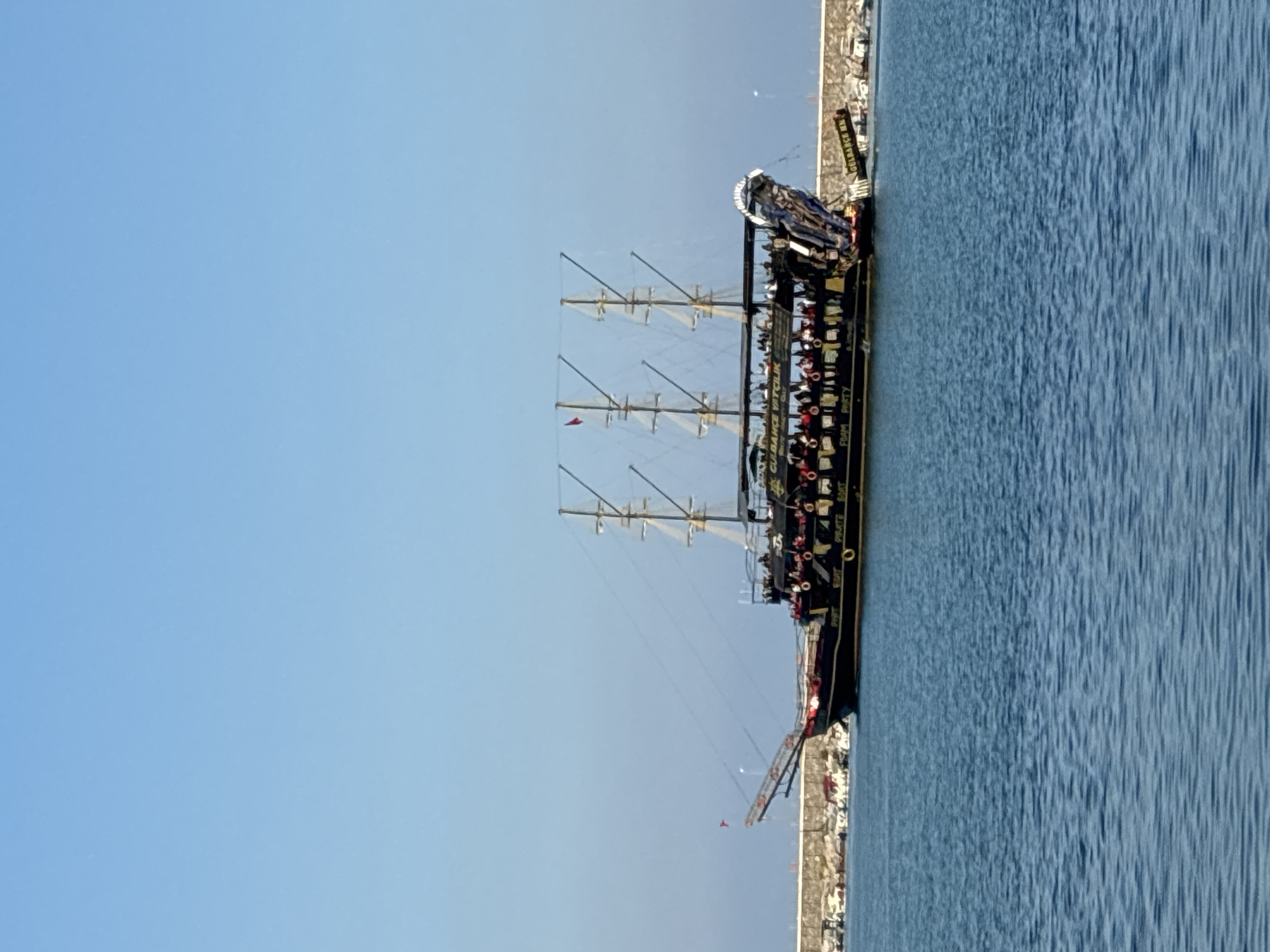 A pirate-themed boat, identified by the text "GULBANCE VATCILIK PIRATE BOAT" visible on its hull, is docked in the blue water of Alanya, Turkey, near a stone breakwater. The boat is painted black with gold accents and features three masts with sails furled. Numerous people are visible on the boat's decks, many wearing red life vests, suggesting a tour or excursion. The boat has a decorative, sculpted bow resembling a dragon or mythical creature. In the background, a stone jetty with scattered smaller boats and debris is visible. The sky is clear and blue.