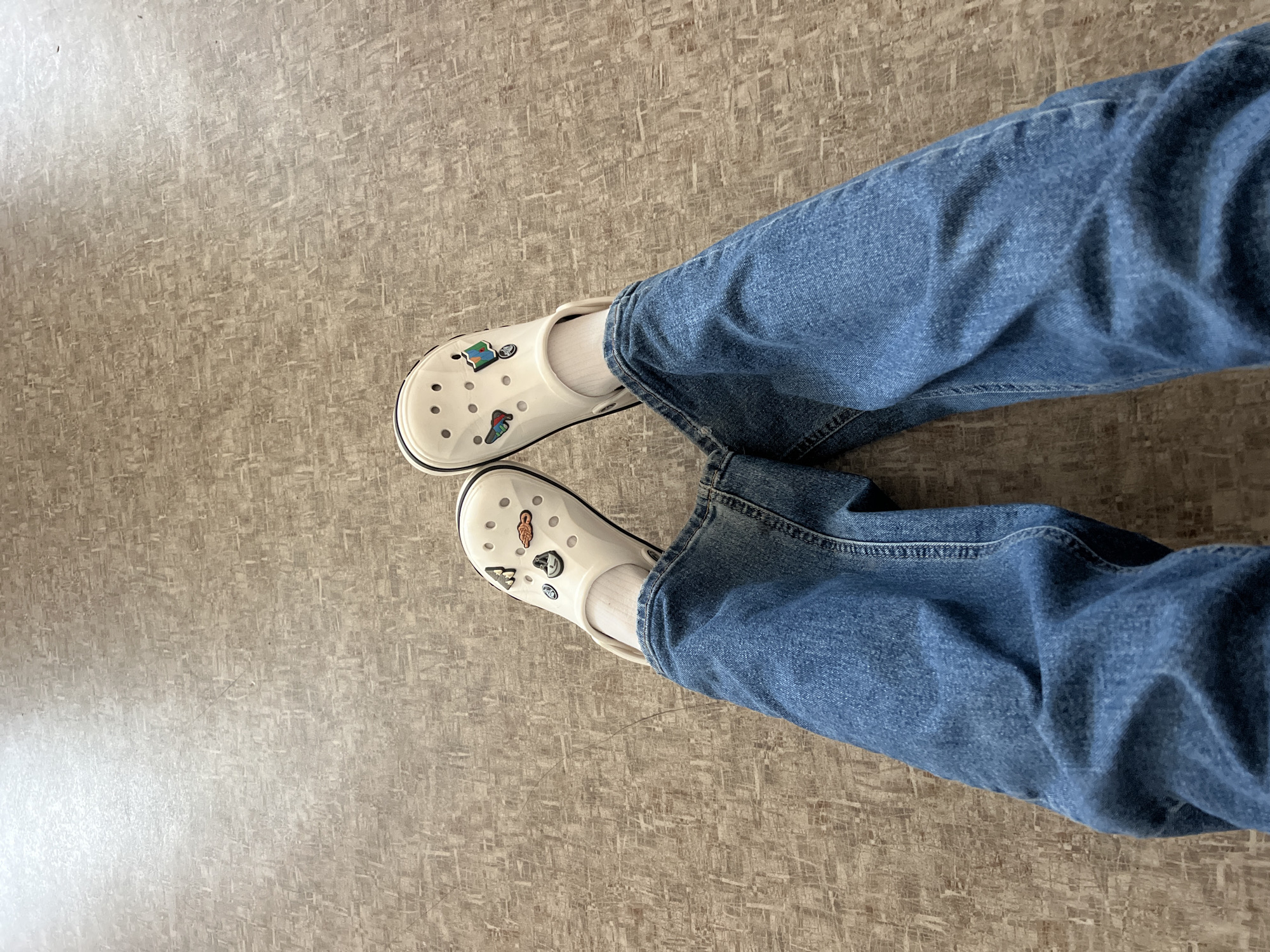 The image shows a person's legs from the knees down, wearing blue jeans and white Crocs with charms. The person is standing on a beige floor with a textured, non-descript pattern. The location is not identifiable as Chaikovsky, Russia from the image alone. No actions or interactions are occurring in the scene.