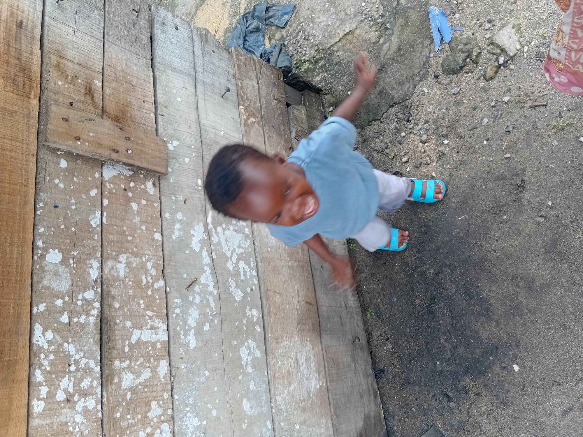 A young child, with a light blue shirt and light-colored pants, is captured in motion near a weathered wooden wall. The child's mouth is open in what appears to be a laugh or shout, and their arms are outstretched. They are wearing bright blue sandals. The wooden wall is constructed of horizontal planks with a weathered, aged appearance, showing signs of wear and tear, with some white splotches and exposed nails. The ground is a mix of dirt and gravel. To the right of the child, there's a pile of dark fabric and some rocky terrain. The scene is outdoors, suggesting a rural or undeveloped urban setting.