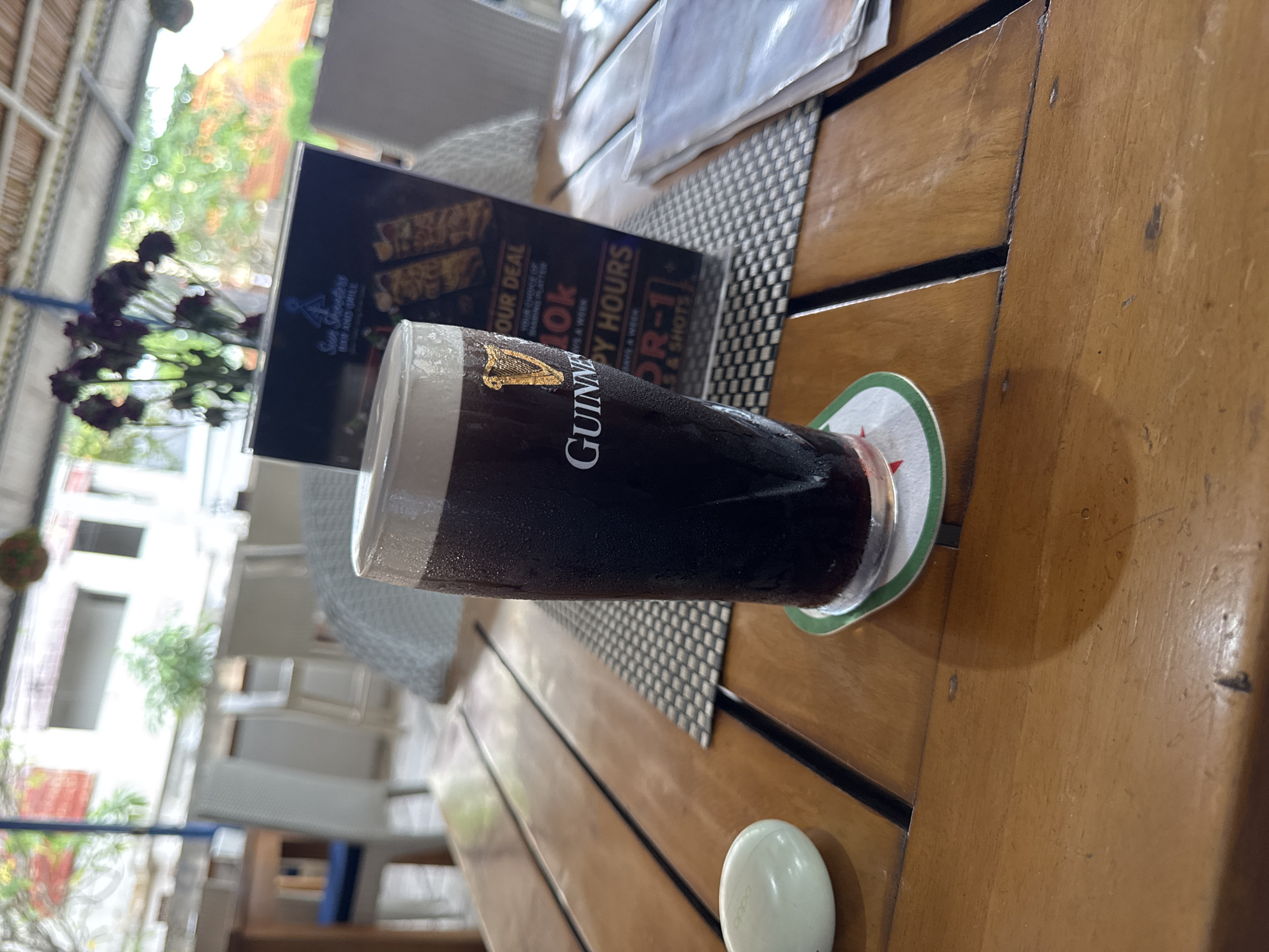 A tall glass of dark beer, identified as Guinness by the logo on the glass, sits on a wooden table. The glass is condensation-covered and has a frothy head. A beer mat is under the glass. Next to the glass is a menu stand advertising "OUR DEAL 10k HAPPY HOURS 2 FOR 1". In the background, there are white chairs and tables, and foliage visible through a covered outdoor seating area that appears to be part of a restaurant or bar. The overall setting suggests a casual outdoor dining or drinking establishment, likely in a tropical location given the lush greenery. The "North Kuta, Indonesia" location tag implies this establishment is situated in that area. There are no people clearly visible in the image.