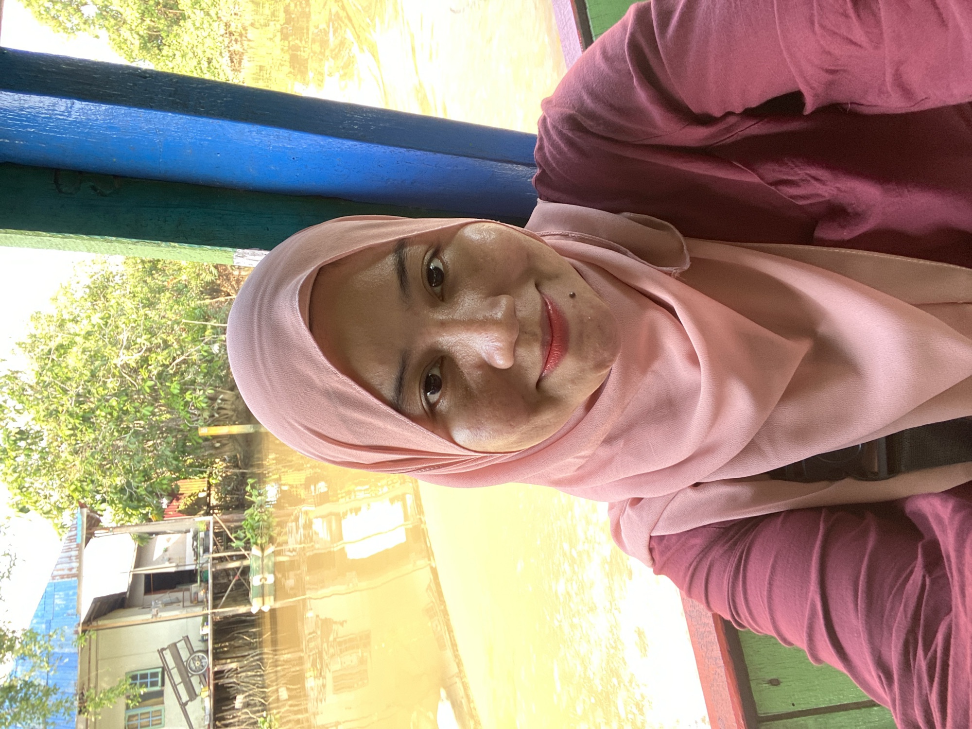 A young woman wearing a pink hijab and a maroon shirt takes a selfie from a boat. She is smiling at the camera. Behind her, the muddy brown water of the Martapura River reflects the surrounding greenery and a stilted house on the bank. The house appears to be made of wood and has a blue corrugated metal roof. A wooden cart with two wheels is visible near the house. The boat's interior is visible with green and red painted wooden railings. The scene is set in Banjarmasin, Indonesia, known for its extensive river system and floating markets.