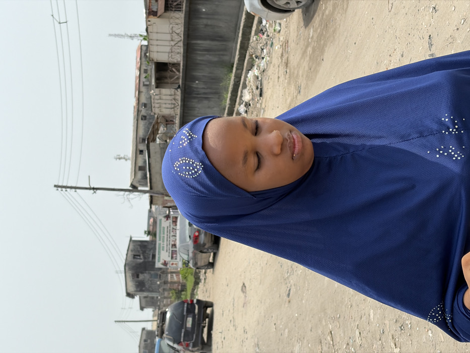 A young individual, likely female, stands on an unpaved dirt road in Jakande, Nigeria, under a bright, hazy sky. She wears a deep blue hijab, which covers her head and drapes over her shoulders and chest, adorned with silver or white rhinestone patterns including an oval-like design and a heart-like shape on the forehead area, and scattered dots on the lower front. Her eyes are closed or cast downwards, and a subtle nose piercing is visible on her left nostril.
The immediate foreground consists of light brown, dusty ground. In the background, an unpaved road extends, with several vehicles parked along its left side, including a dark SUV and a light-colored sedan. Further right, a partial view of a light-colored car's front bumper is visible. A large, faded billboard or sign with text and images stands between the vehicles and distant structures. Multiple utility poles with numerous overhead power lines traverse the scene horizontally. Along the right side of the road, a concrete wall topped with barbed wire separates the road from accumulated trash and debris. Behind this wall, and further in the distance, are several dilapidated, multi-story buildings under the hazy sky.