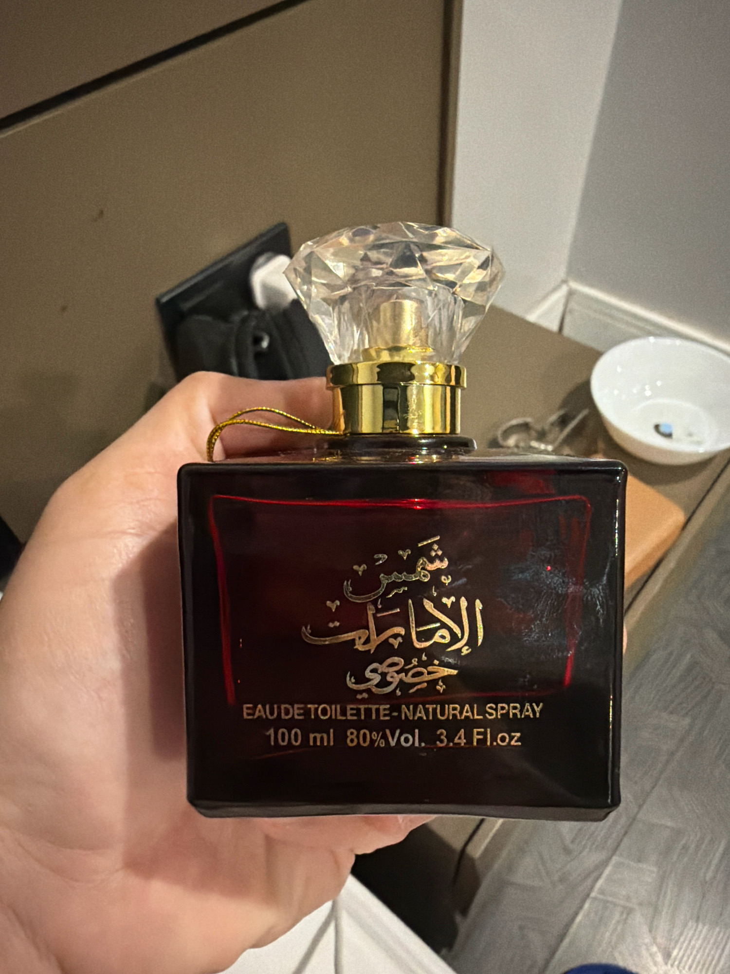 A hand is holding a rectangular, dark red perfume bottle with a clear, faceted cap. The bottle has gold Arabic lettering on the front, which reads "شمس الأمراء" (Shams Al-Umara). Below the Arabic text, it says "EAU DE TOILETTE - NATURAL SPRAY 100 ml 80% Vol. 3.4 Fl.oz". A thin gold string is attached to the neck of the bottle. In the background, there is a brown wall, a white bowl, and a black object that appears to be a bag or wallet. The overall lighting suggests an indoor environment. The image was taken in Ataşehir, Turkey.