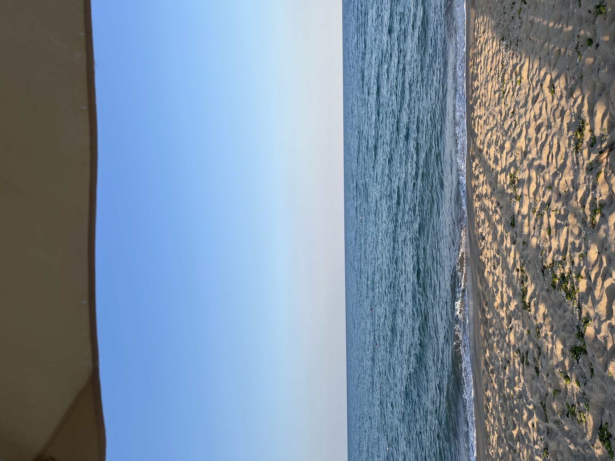 This is a vertical, landscape-oriented photograph taken at a beach, likely during the day given the bright sunlight casting shadows. The scene is divided into three distinct bands. On the far left, a portion of a beige umbrella or awning is visible, with its curved edge creating a partial frame for the image. To the right of the umbrella, a vast expanse of clear, light blue sky dominates the upper portion of the frame, transitioning to a paler, almost white hue closer to the horizon. The middle section of the image features the calm, blue-green ocean, with gentle waves lapping at the shore. The water's surface is textured, showing the ripples and movement of the sea. The rightmost section of the photograph showcases the sandy beach. The sand is light brown and marked with footprints and the texture of dry sand. Shadows from an unseen object, likely the umbrella or a nearby structure, create a pattern of diagonal lines across the sand. Small patches of green vegetation are scattered on the sand. The overall impression is one of a peaceful and serene beach scene, typical of a sunny day at the coast. There is no visible text in the image.