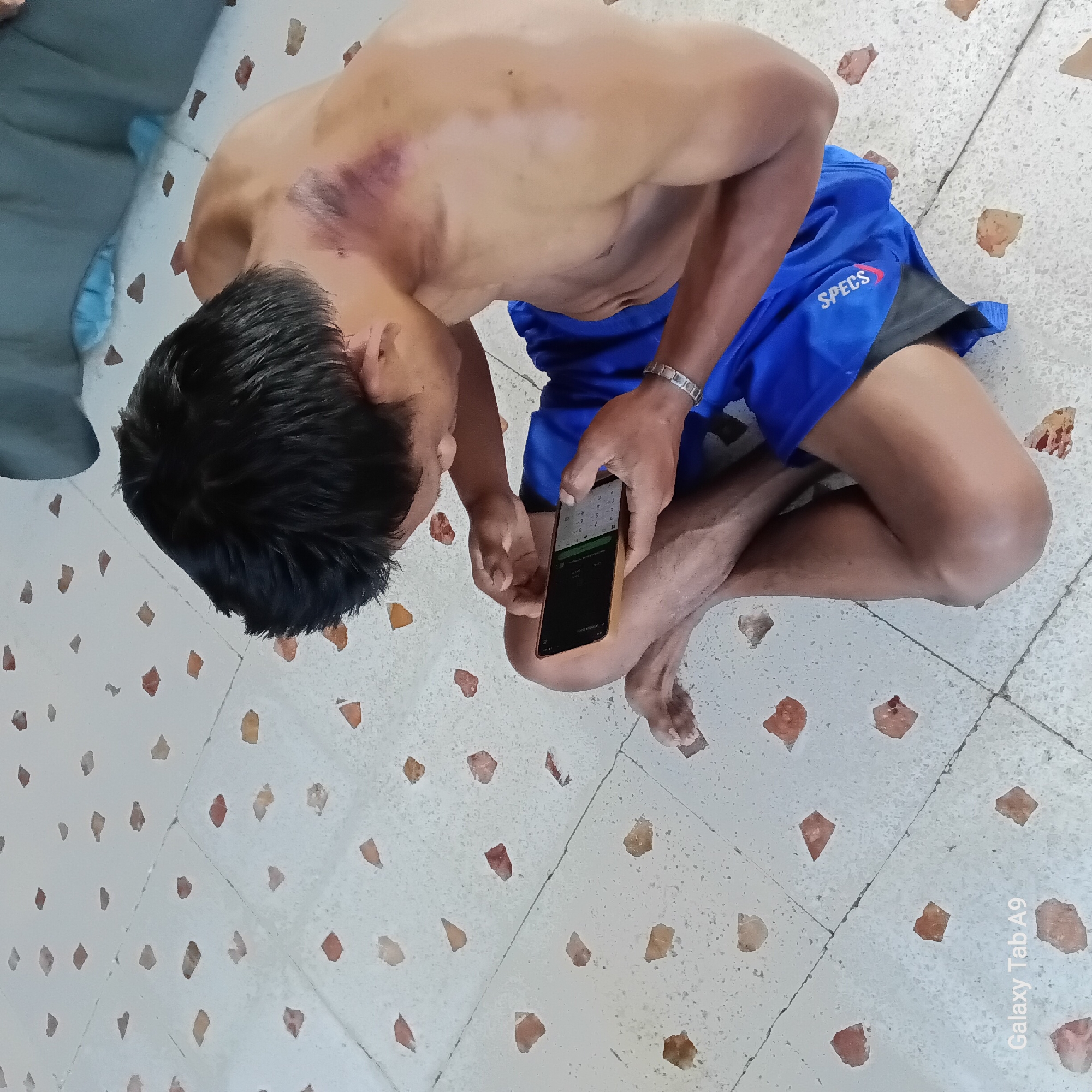 The image shows a person sitting on a tiled floor, holding a smartphone. The person is shirtless, and there is a visible bruise on their upper chest and shoulder area. They are wearing blue athletic shorts with the word "SPECS" printed in white on the left leg. The person is looking down at their phone, which displays a keypad or similar interface. The floor is covered in light-colored tiles with scattered reddish-brown inserts. The lighting suggests it might be daytime, possibly indoors or in a shaded outdoor area. The overall mood is neutral, with the focus on the person and their interaction with the phone. The visible text on the image is "SPECS" on the shorts and "Galaxy Tab A9" in the bottom right corner. The location context is Sambirejo, Indonesia.