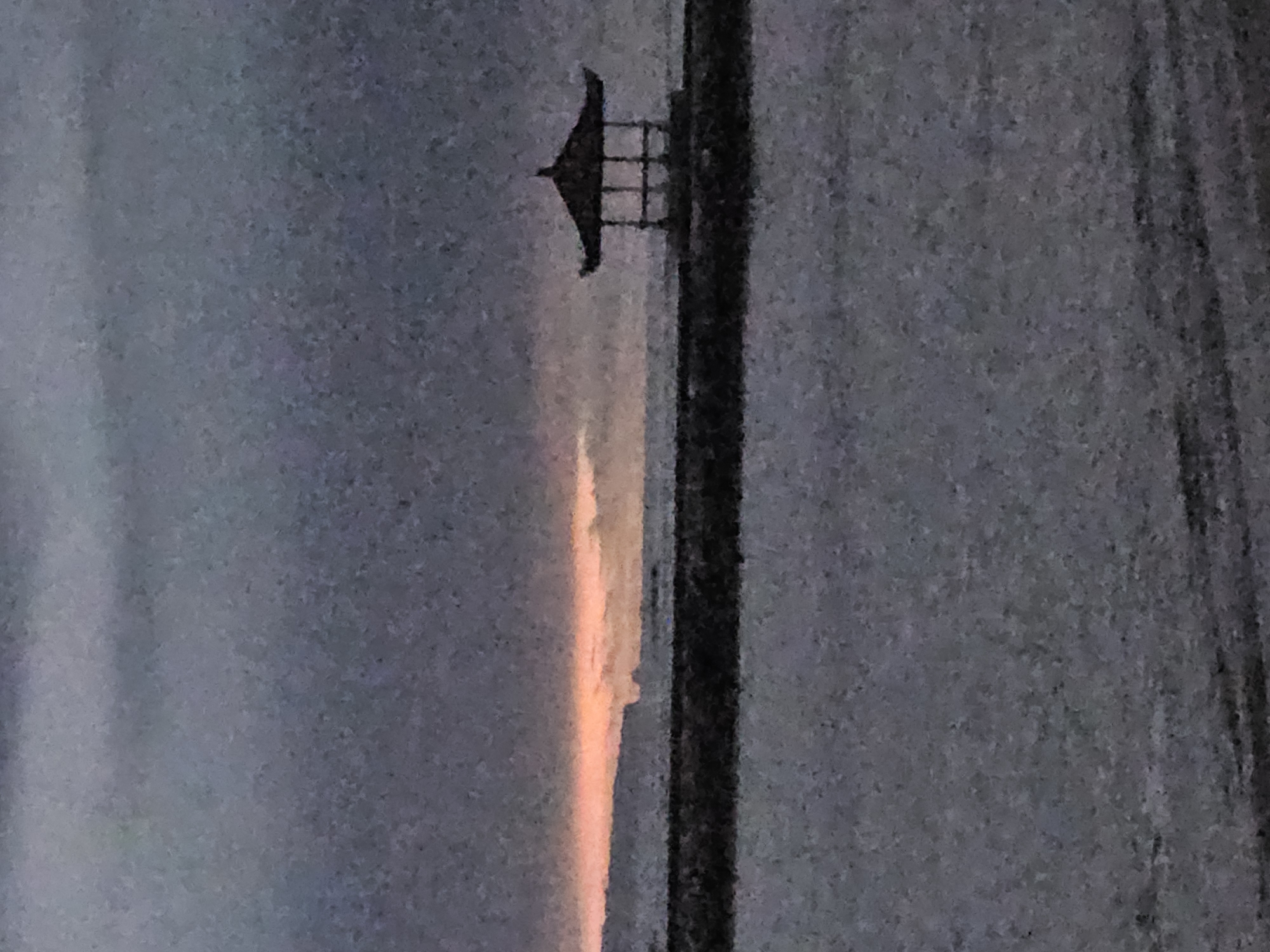 An obscured image of a utility pole with a weather vane, taken vertically. A sunset is visible in the background. The location may be Denpasar, Indonesia.