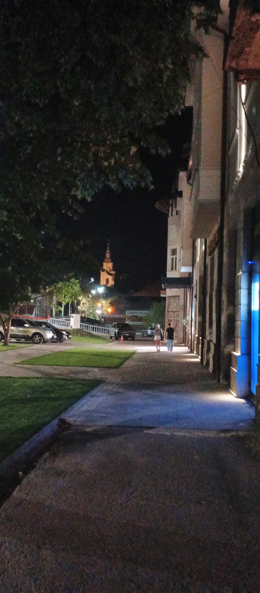 The image depicts a nighttime scene on a city street in Berehove, Ukraine, likely in the downtown area. A large tree with dark foliage dominates the left side of the frame, casting shadows. To the right, a multi-story building with a stone facade extends along the sidewalk. The building features tall, narrow windows and a row of vertical architectural elements. A bright blue light emanates from an opening in the building on the far right.
A paved sidewalk runs diagonally from the foreground towards the background. Two individuals, a man and a woman, are walking away from the camera on this sidewalk, illuminated by streetlights. The woman is wearing a light-colored top and skirt, while the man is in a dark shirt and pants.
Across the street, a grassy area is visible, with several parked cars, including a silver sedan and a dark SUV. Beyond the grassy area, there's a lower building with a sign that appears to be in Cyrillic. Further in the distance, a prominent church tower with a pointed spire is visible, illuminated by lights. Streetlights are scattered throughout the scene, providing illumination and creating pools of light on the pavement. The overall atmosphere is that of a quiet urban evening.