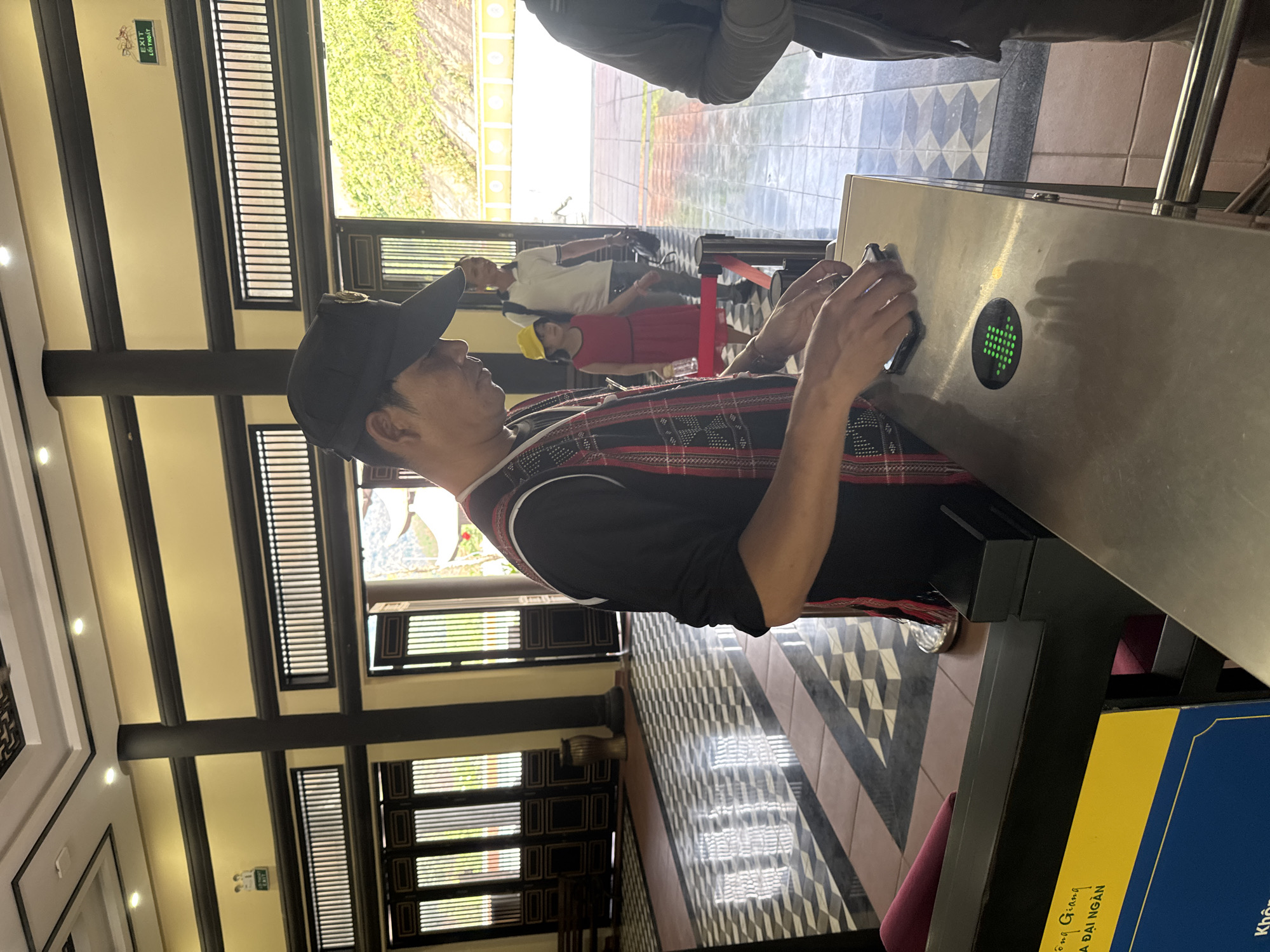 An indoor scene depicts people at what appears to be an entry or exit point, likely a turnstile, in Đà Nẵng, Vietnam. The primary subject is a man, viewed from the side, wearing a black cap, a black t-shirt with a red and white patterned design on the shoulders and upper back, and dark pants. He is holding a small electronic device, possibly a smartphone or ticket, over a silver turnstile unit with a bright green arrow indicator, suggesting a successful scan or permission to proceed.
In the background, behind the main subject, another man in a white polo shirt and a woman in a red dress with a yellow hat are waiting. Beyond them, a checkered black and white floor transitions to a lighter, possibly tiled or paved, outdoor area visible through large windows. The interior features a decorative ceiling with dark wooden beams and light-colored panels, illuminated by recessed spotlights. Windows with horizontal slats allow natural light into the space, showing greenery outside.
Notable architectural details include the dark wooden beams and intricate window designs, giving the building a traditional or styled appearance. The floor features a repeating diamond and square pattern in black, white, and grey tones.
Visible text includes an "EXIT LỐI THOÁT" sign (Vietnamese for Exit) on a green plaque on the wall. A blue sign near the turnstile has partially visible Vietnamese text: "...Sông Giang ĐÀI NGÀN...", which might refer to a specific location or organization. The bright lighting suggests it is daytime. The overall atmosphere is functional, with people moving through a controlled access point.