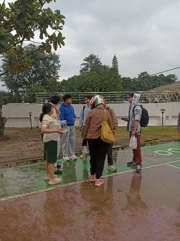 Six people are gathered in an open area in Jatinangor, Indonesia, at the coordinates -6.931 latitude, 107.776 longitude. The ground is wet and appears to be a paved area with some green grass and trees surrounding it. One person is wearing a motorcycle helmet. Two people are holding bags. Two people are wearing backpacks. Five are wearing casual attire, one person is wearing a hoodie with a pattern and a scarf. The person on the left is holding a white object in their left hand. There is a railing in the background, and some foliage behind them. The overcast sky appears cloudy. The individuals are engaging in a conversation, as indicated by their stances and the direction they are facing. They appear to be interacting with each other. The environment is calm and relaxed.