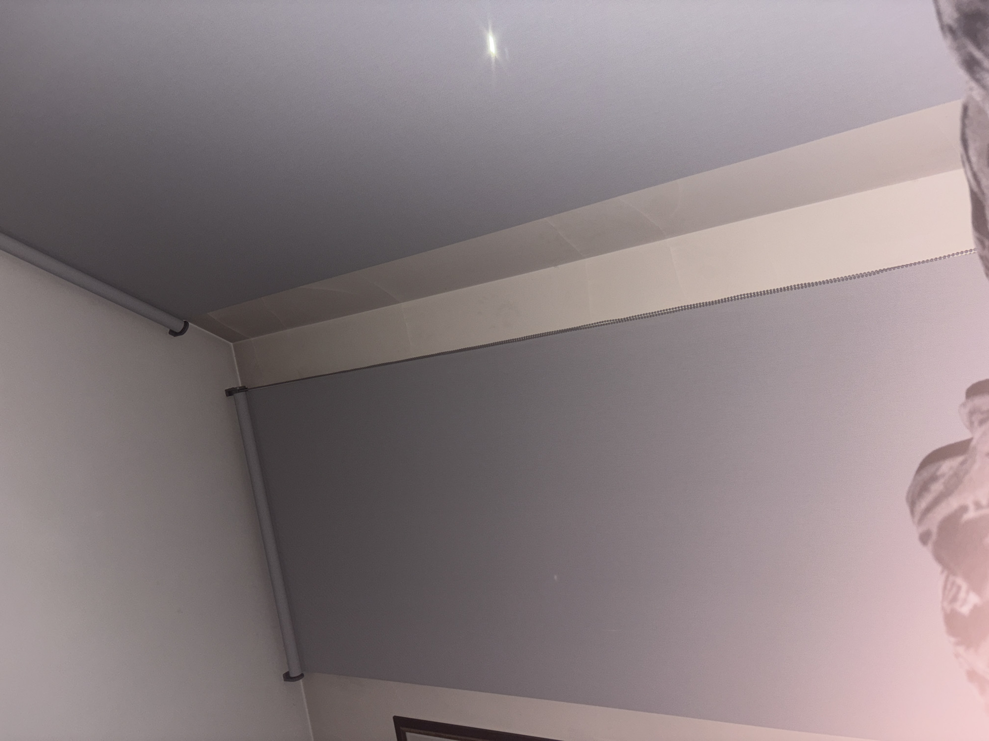 The image depicts an indoor scene in Plovdiv, Bulgaria, viewed from a low angle looking upwards into a room corner. The ceiling is white. The walls are a uniform light grey-purple color. A grey roller blind is mounted near the ceiling along the left and central wall, secured by black brackets. A metallic bead chain, used to operate the roller blind, hangs vertically along the right side of a prominent, lighter-toned vertical structural element or panel on the right wall. This lighter panel features a vertical seam or shadow line, suggesting it is composed of two narrower sections. A bright, elongated reflection is visible on the upper right portion of the grey-purple wall. On the far left, a dark-framed object, possibly a mirror or picture frame, is partially visible on an adjacent wall section. The bottom foreground of the image shows blurred, crumpled fabric, consistent with bedding or soft furnishings.