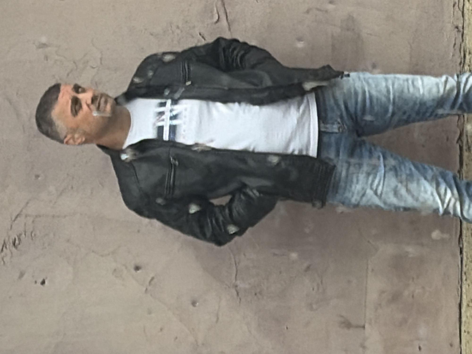 A man stands against a light brown stucco wall with dark spots and a vertical wooden board along the right edge. He is wearing a black leather jacket over a white t-shirt with a navy blue design, and light blue jeans. A cigarette is held between his lips. The location is Sofia, Bulgaria.