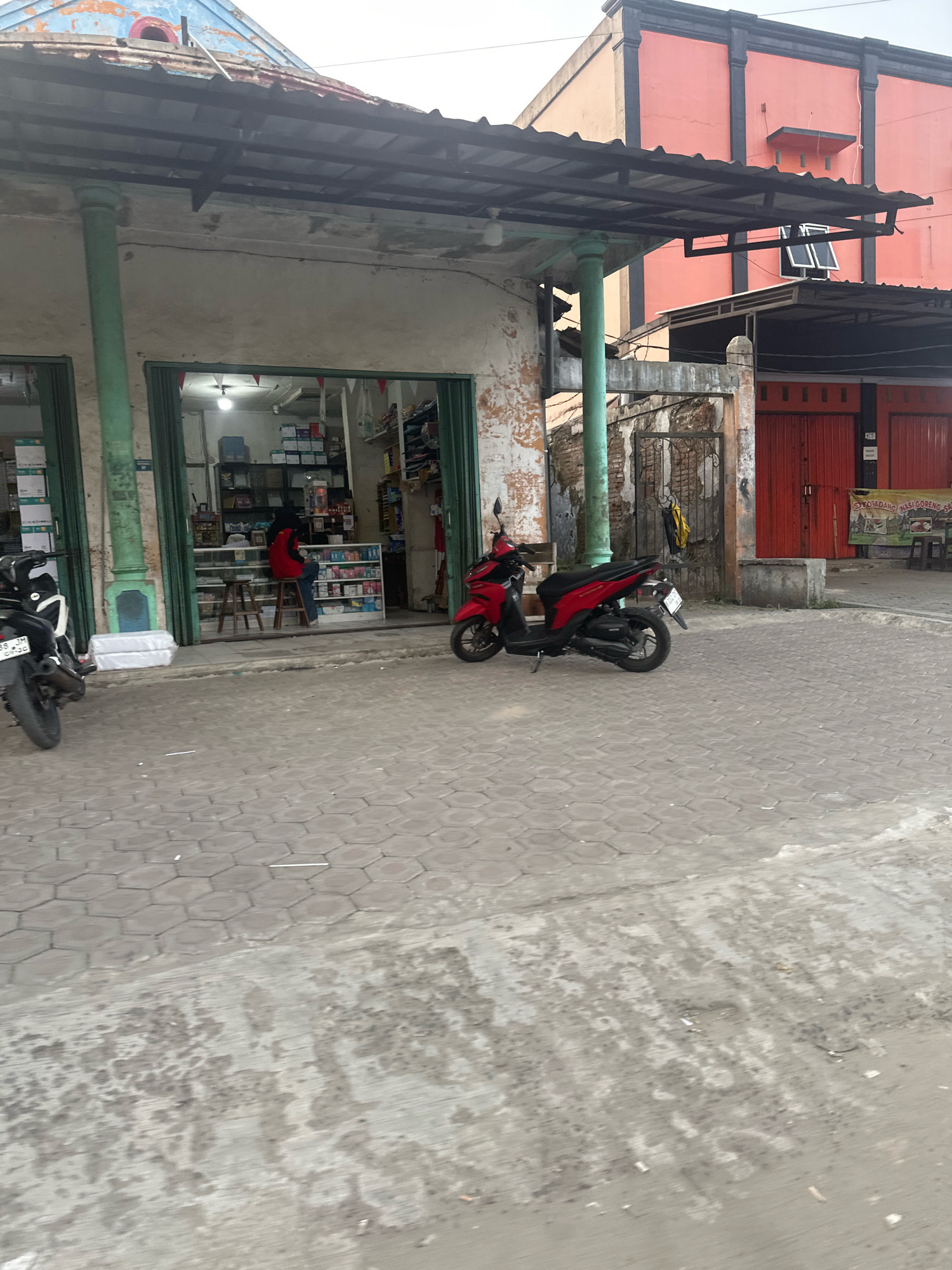 The image captures a street scene in Karaton, Indonesia, focusing on a small shop with an open storefront. A red and black scooter is parked prominently in front of the shop, facing left. To the left of the scooter, a black and white motorcycle is partially visible. Inside the shop, shelves are stocked with various items, and a person wearing a red jacket and blue jeans is seated on a stool. The shop's facade is weathered, with peeling paint on the roof and walls. In the background, an orange building with dark trim stands out. The street is paved with hexagonal interlocking blocks, and a section of the foreground shows a worn concrete surface with some patchy discoloration. The overall atmosphere suggests a typical day in a local commercial area.