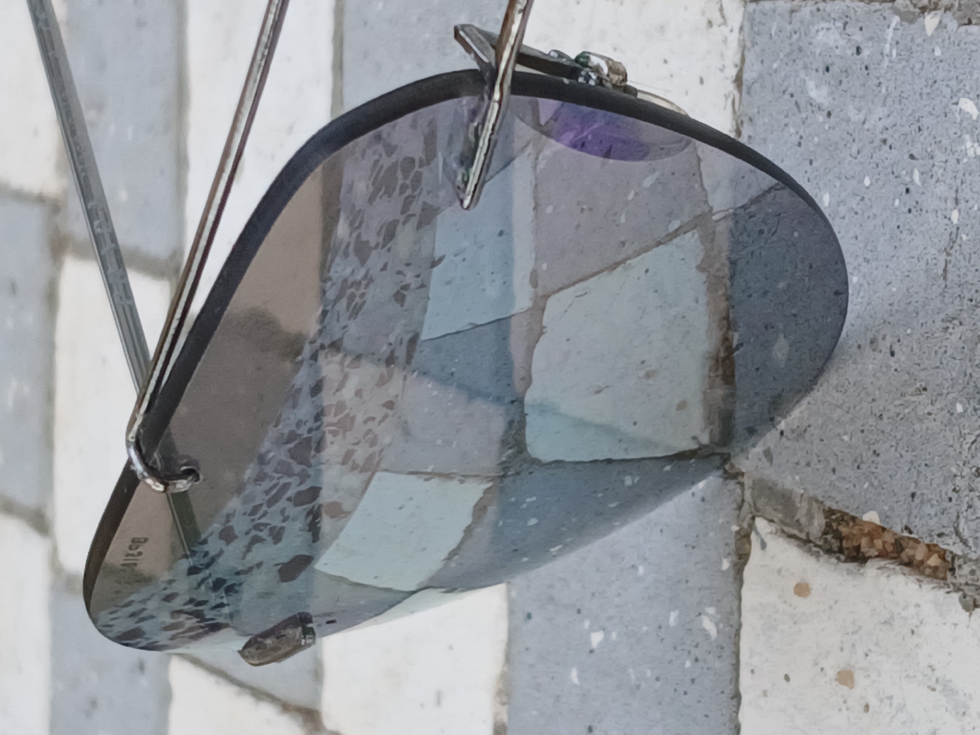 The image displays a single, dark-tinted lens, likely belonging to sunglasses or rimless eyewear, resting on an outdoor tiled surface. No people are visible in the frame.
The lens itself is oval-shaped and appears to be made of a synthetic material. Its surface exhibits a mottled texture, possibly from water spots, smudges, or a specific coating, giving it a somewhat irregular transparency. Reflections of the grey and light-colored square and rectangular tiles beneath are visible on the lens. A faint purple hue can be seen in some sections of the lens, particularly towards the top right.
Metallic frame components are attached to the top of the lens, with thin, silver-colored wires extending upwards, suggesting these are the arms or part of the bridge of the eyewear. On the lower left portion of the lens, white text is visible, clearly spelling "Boss" with a stylized "o" (a small circle with a dot inside) and unique "ss" characters.
The setting appears to be an outdoor paved area, possibly a patio or sidewalk, composed of irregularly shaped and arranged grey and light-colored concrete or stone tiles. The grout lines between the tiles show some wear and minor debris. The lighting suggests it is daytime, with soft, diffused light, as there are no harsh shadows. No specific activity is occurring; the lens seems to be stationary, perhaps having been placed or dropped. There are no direct location cues within the image that specifically point to Ciudad Guayana, Venezuela, beyond the general appearance of an outdoor paved area.