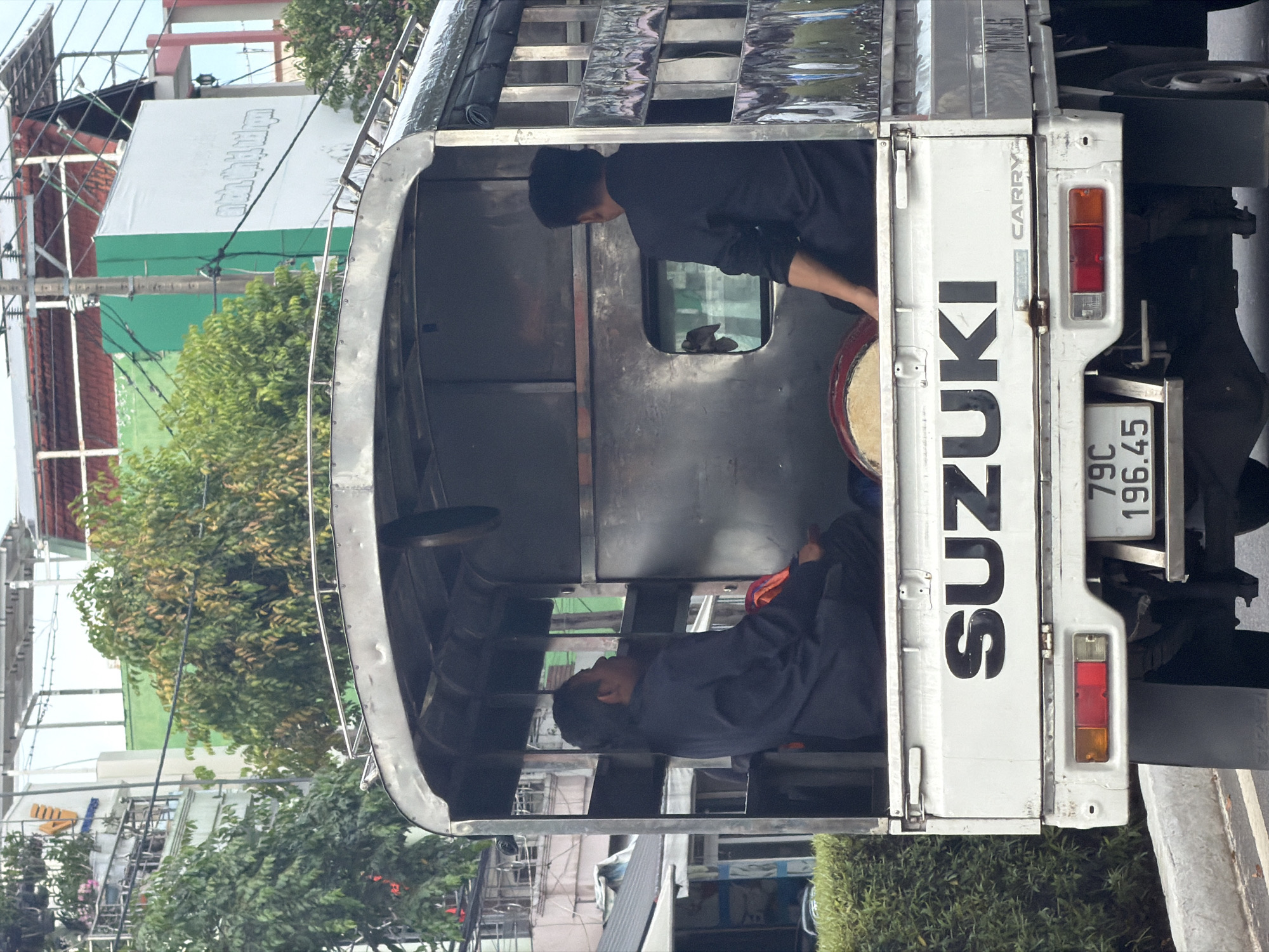 The image shows a white Suzuki Carry truck parked on the side of a street. Two people are visible in the back of the truck, one sitting and one standing near the door with a drum. The license plate reads "79C 196.45." The photo was taken in Tay Nha Trang Ward, Vietnam, and the background includes buildings, power lines, and trees.