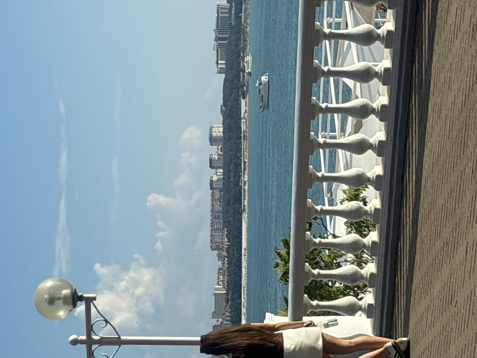 The image depicts a sunny day with a clear blue sky and scattered clouds. In the foreground, a woman with long dark hair is walking away from the camera, wearing a dark top and a white skirt, and carrying a clear plastic bottle. She is on a paved walkway with a patterned surface, and to her right is a white balustrade with ornate pillars, overlooking a body of water. Beyond the balustrade, a blue sea stretches towards a coastline lined with trees and buildings. Several white yachts are visible on the water. The architecture of the buildings on the coastline suggests a resort or urban area. The scene appears to be a promenade or esplanade in Gelendzhik, Russia, likely overlooking the Black Sea. The overall atmosphere is peaceful and scenic, characteristic of a coastal town.