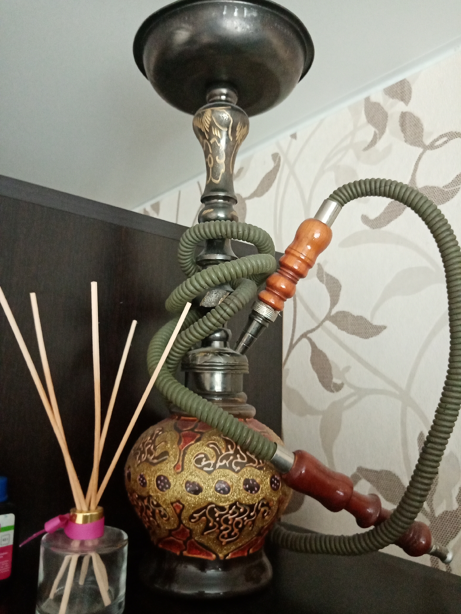 A hookah with a green corrugated hose sits on a dark surface. Next to the hookah is a glass bottle containing several thin wooden sticks, held together by a pink ribbon around the bottle's neck. The background is a wall with a wallpaper pattern of light brown leaves on an off-white background. Elektrostal, Russia.